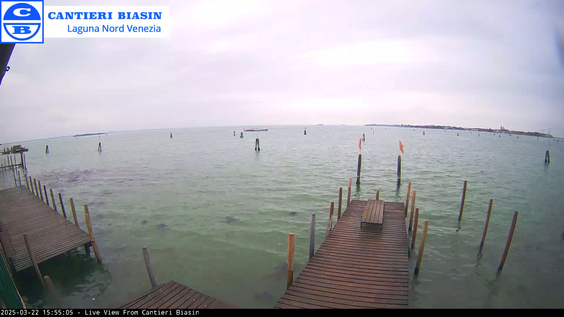 A wooden dock extends over calm, greenish-grey water under an overcast sky, with numerous poles and a small boat visible in the distance.