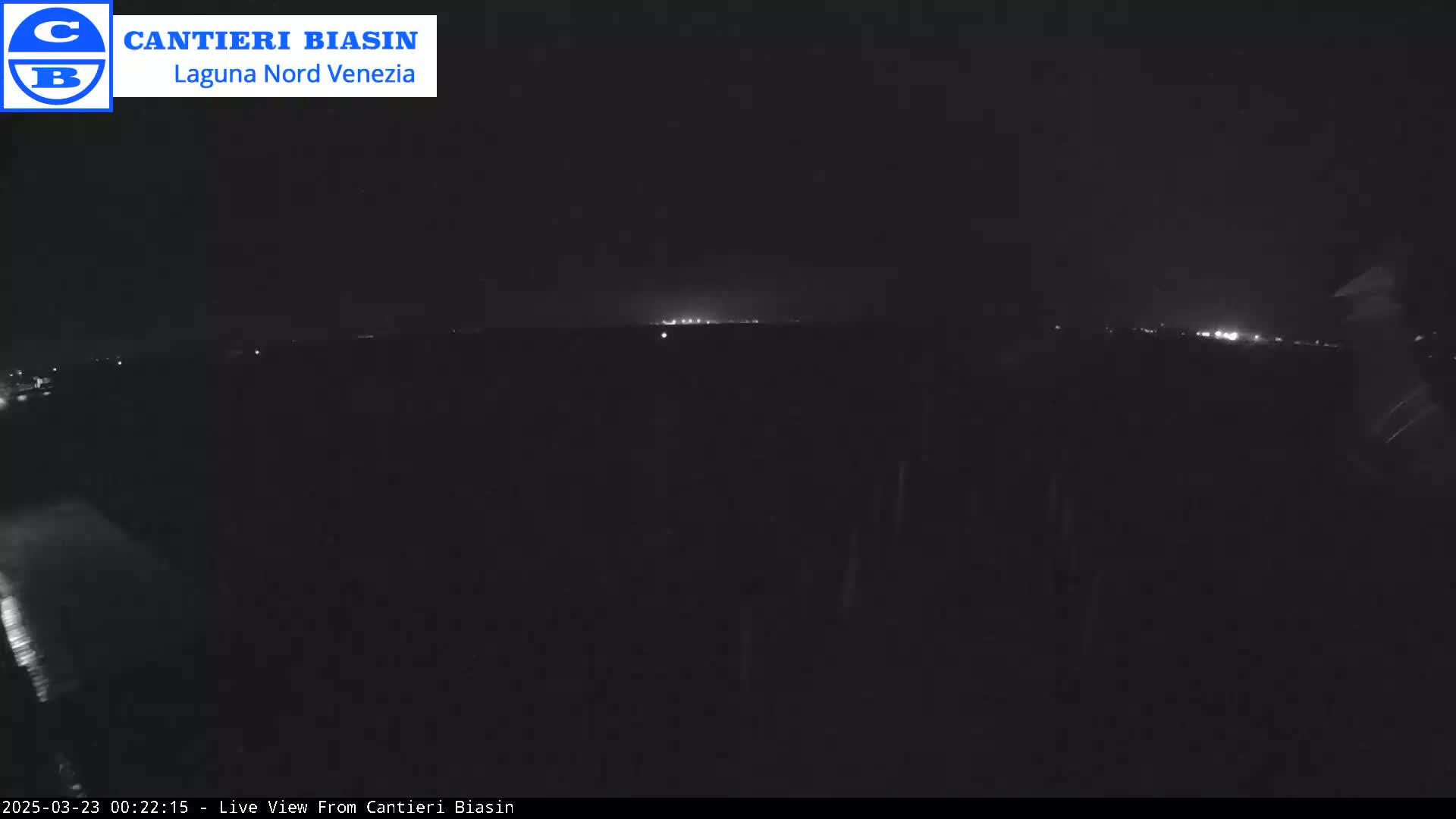 A nighttime, dark outdoor view shows a distant line of faint lights on the horizon, possibly a town, under what appears to be rainy conditions.