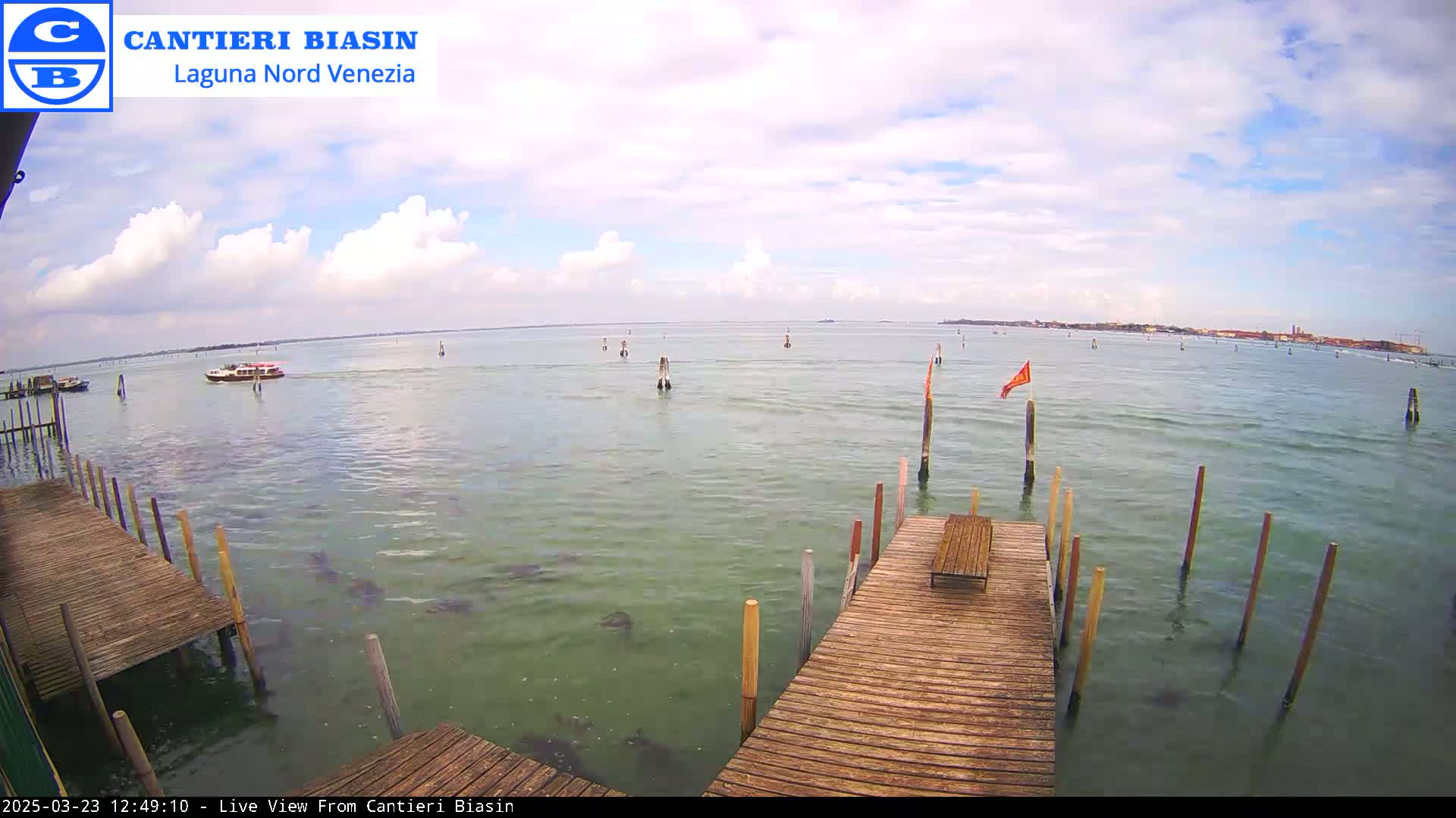 A wooden dock extends over calm, greenish water under a partly cloudy sky, with a small boat visible in the distance and a distant shoreline.