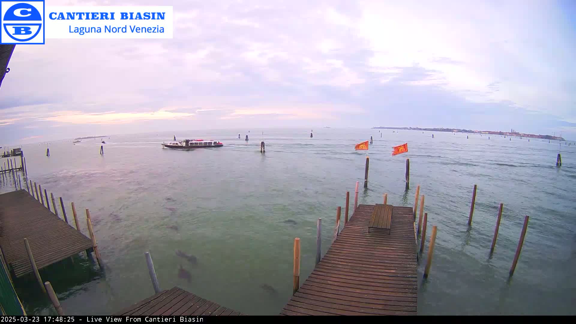 A calm, overcast day shows a body of water with a boat, several posts in the water, and a wooden dock extending from the bottom right corner into the water.