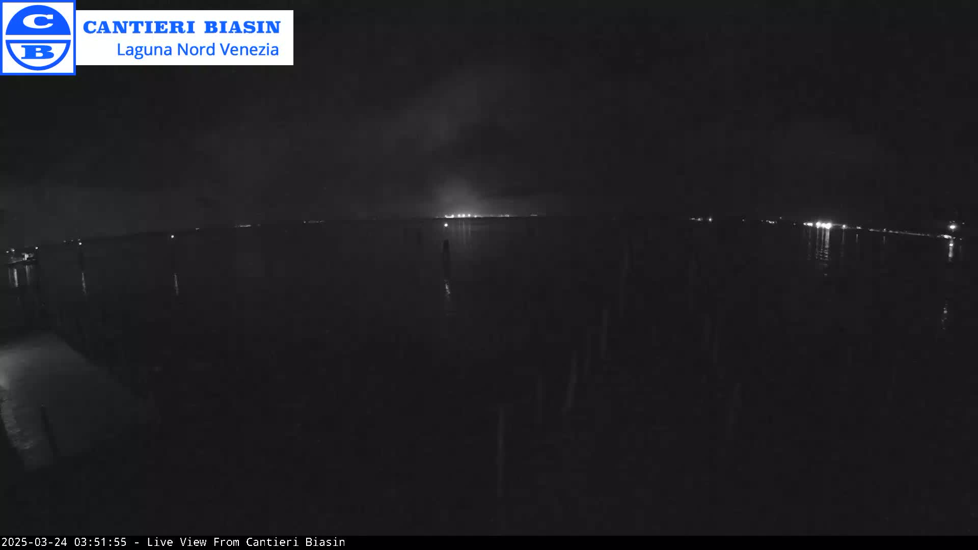 A nighttime view of a dark body of water with faint lights on the opposite shore under a mostly clear sky.