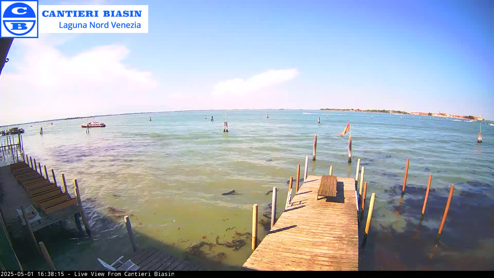 A wooden dock extends over murky green water toward a distant shoreline under a mostly sunny sky, with a small boat visible in the distance.