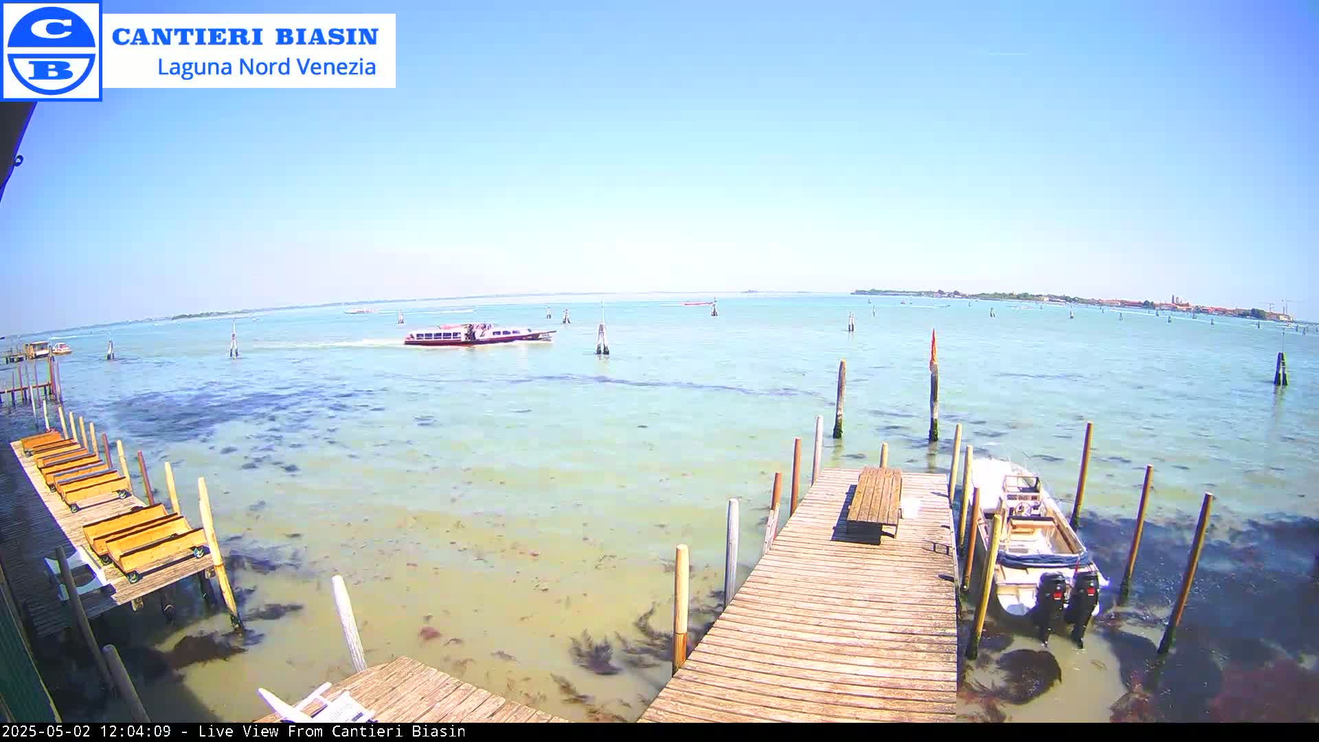 A wooden dock extends over calm, shallow, light-green water, with a boat moored alongside and a passenger boat in the distance on a sunny day.