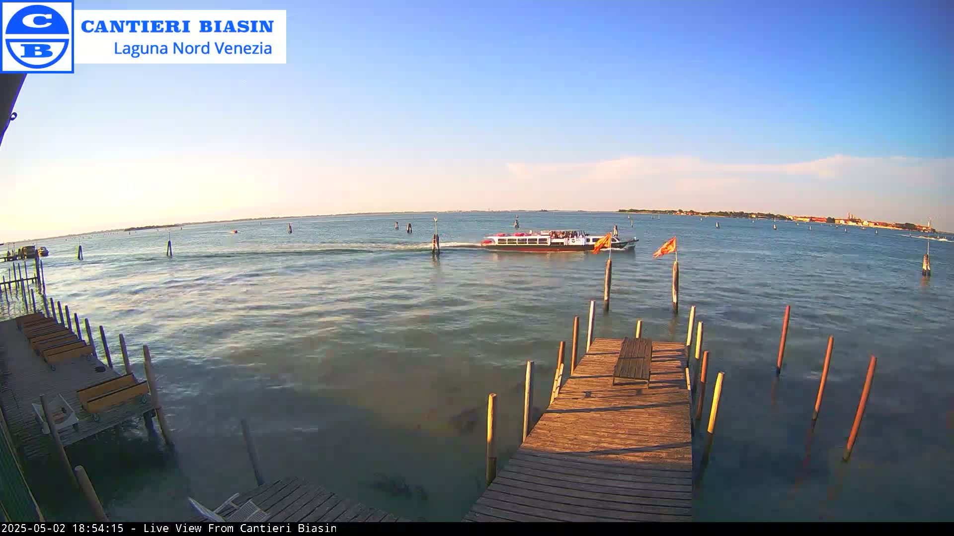 A passenger boat travels across calm, sunlit water toward a distant shoreline, past a wooden dock extending from the foreground.