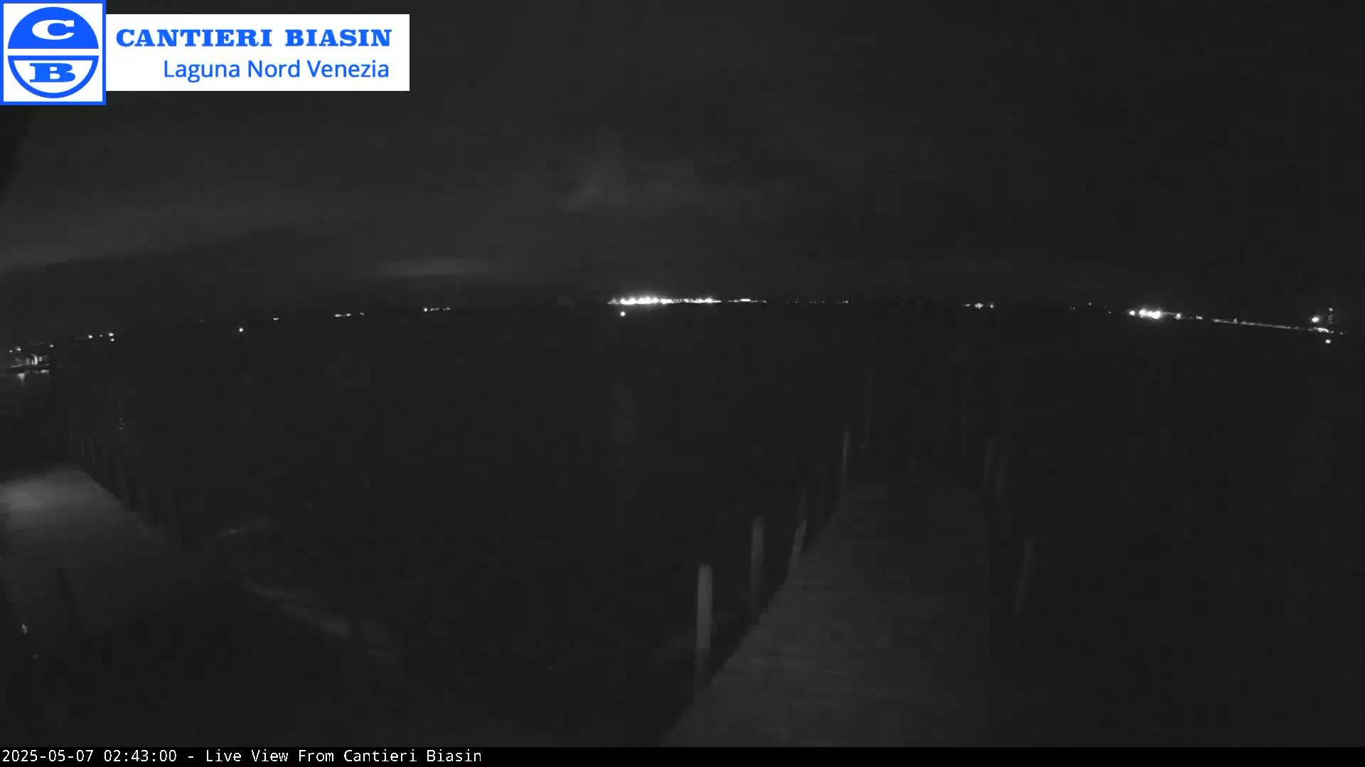 A nighttime, low-light view shows a dark landscape with a distant line of lights, and a wooden pier extending into the foreground.
