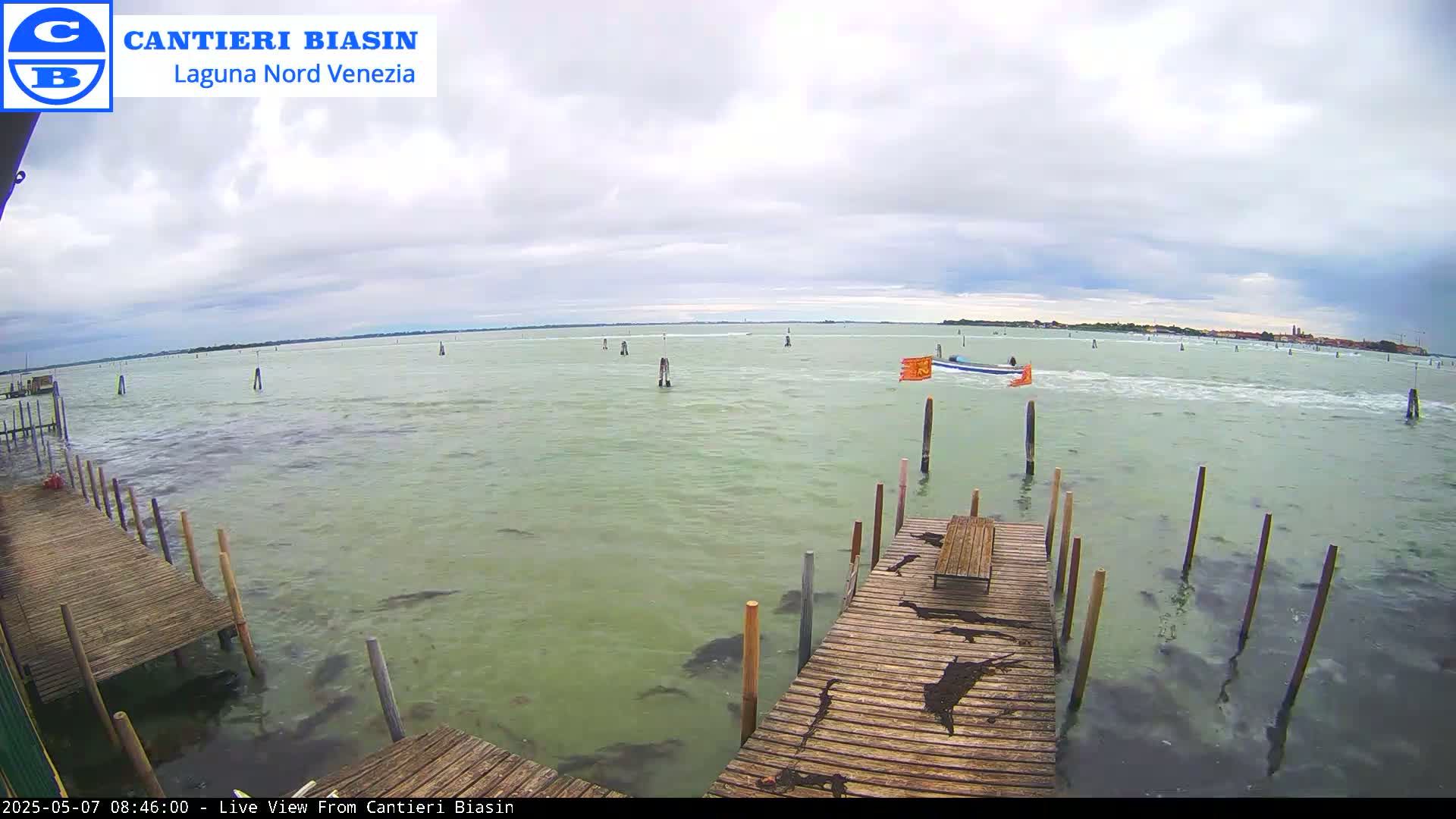 A wooden dock extends over calm, green-tinged water under a cloudy sky, with a small boat visible in the distance.