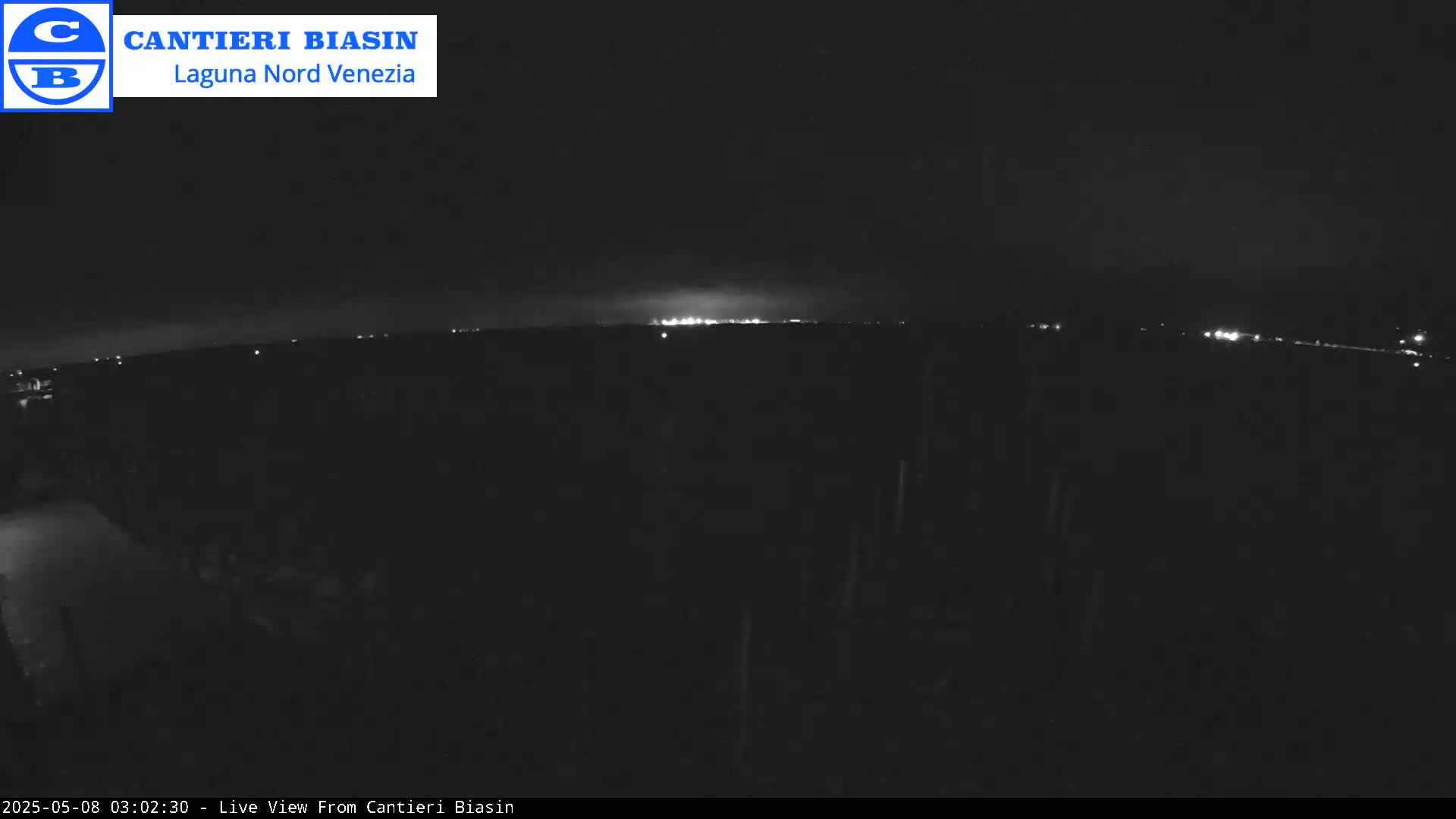 A nighttime, dark view shows a distant line of lights across a flat, dark landscape under a mostly clear sky.