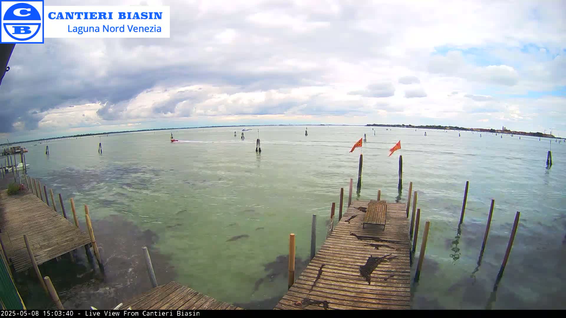 A mostly cloudy day shows a calm body of water with a wooden dock, posts, and small orange flags extending into the water.