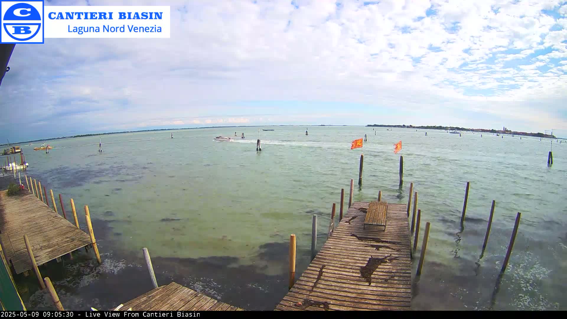 A partly cloudy day shows a calm body of water with several boats, a wooden dock, and distant land.