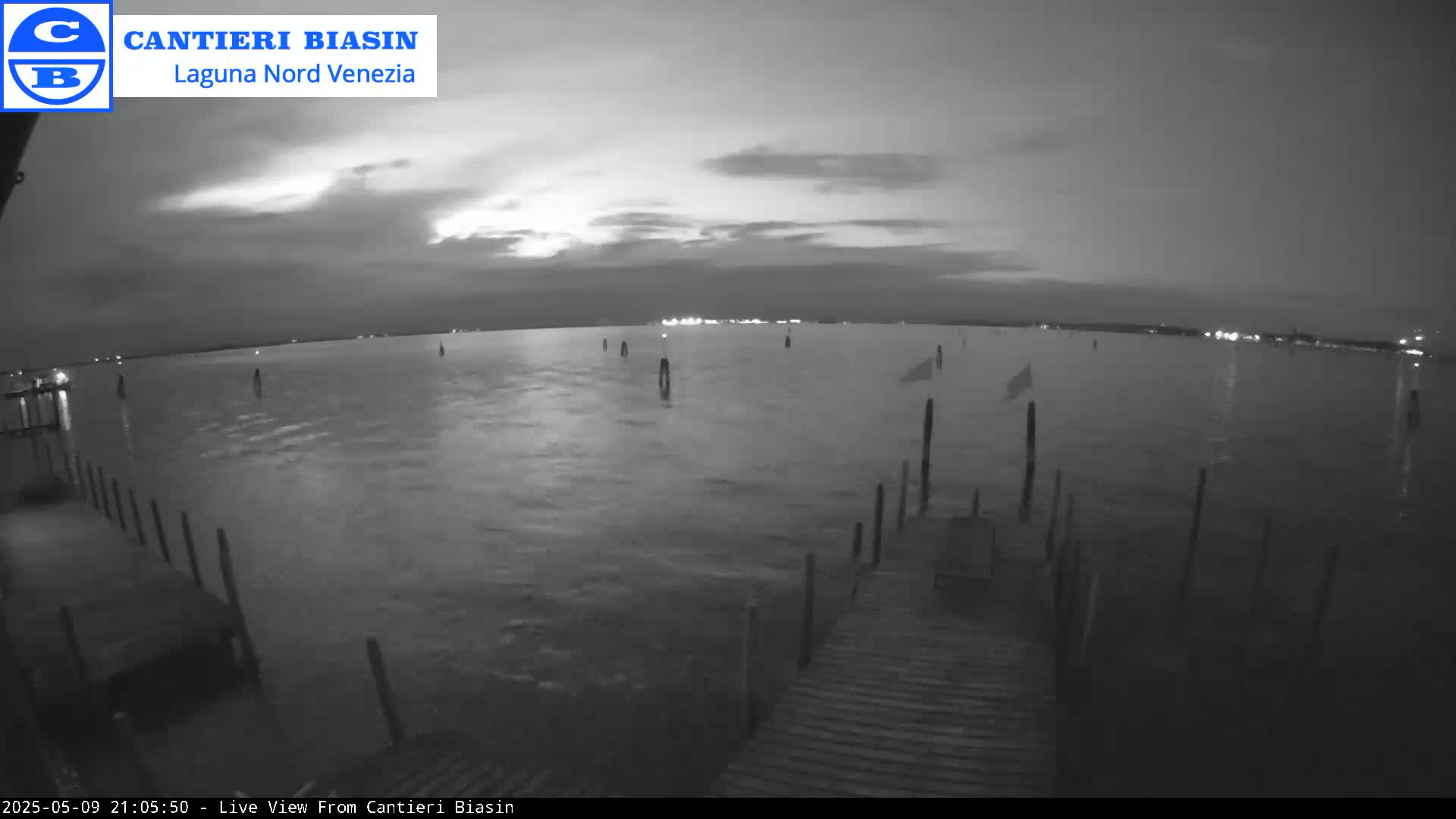 A nighttime view of calm water shows a wooden dock extending into the water with posts in the water, and a distant shoreline with faint lights under a mostly cloudy sky.