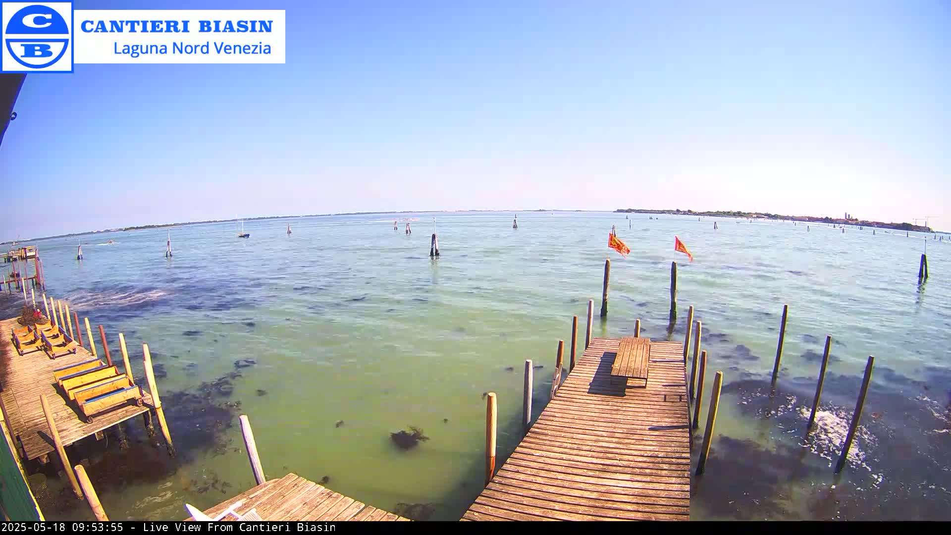 A wooden dock extends over calm, light-green water under a clear blue sky, with a distant shoreline visible.