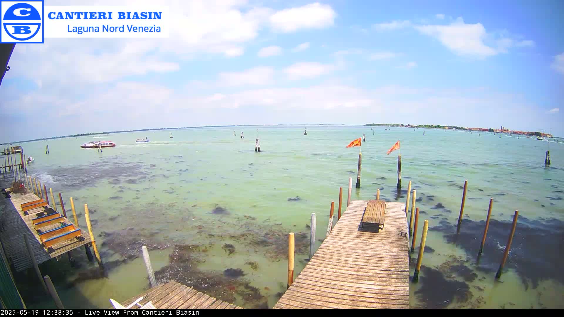 A wooden dock extends over shallow, green-tinged water under a partly cloudy, sunny sky, with boats visible in the distance.