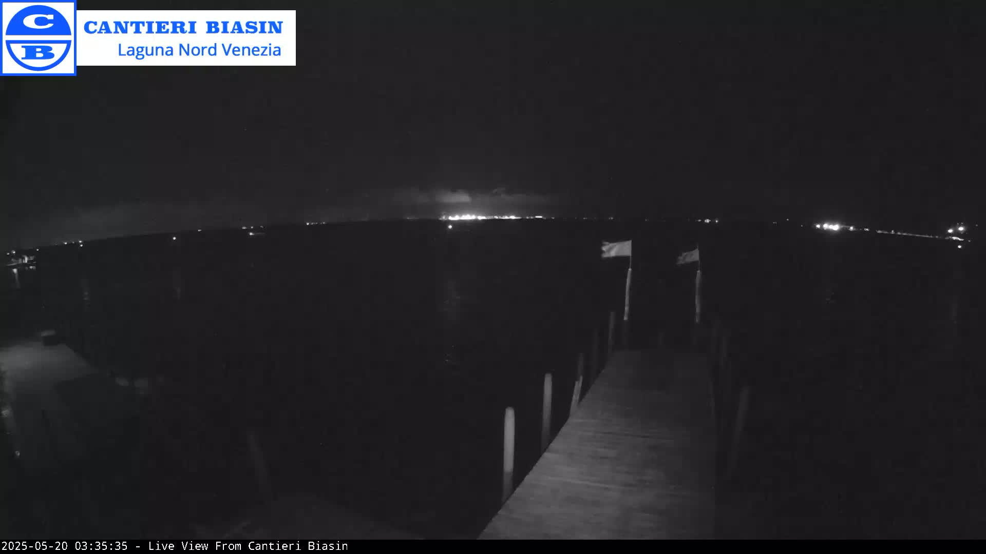 A nighttime view of a wooden dock extending over dark water towards a faintly lit shoreline under a mostly clear sky.