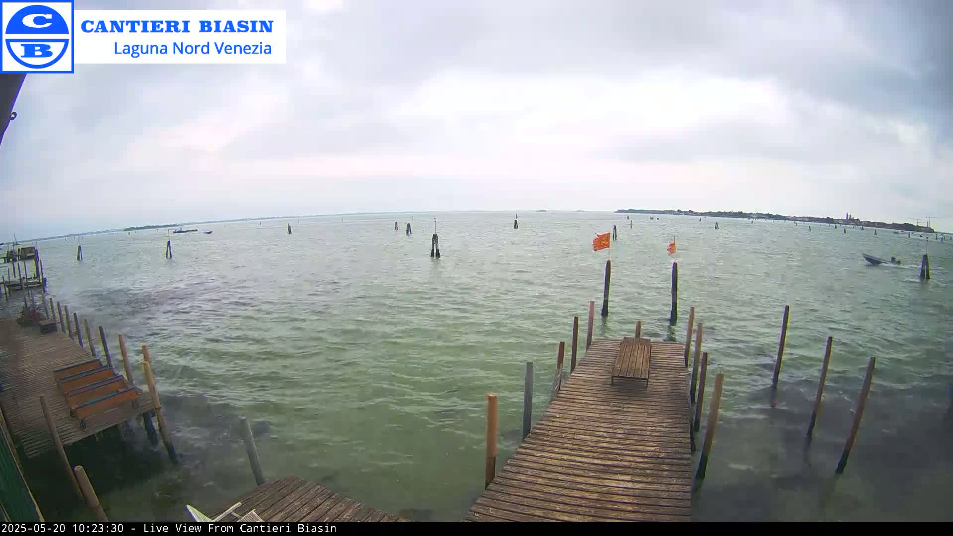 A wooden dock extends over a calm, greenish-gray body of water under an overcast sky, with several posts and small orange flags visible in the distance.