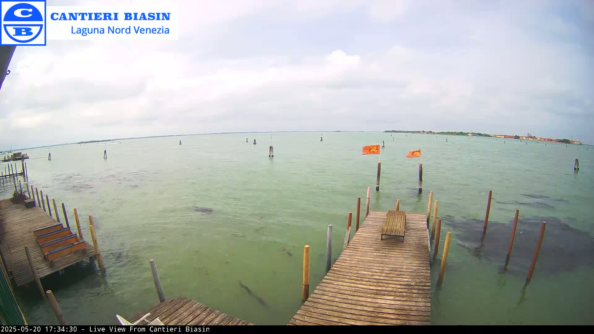 A wooden dock extends over calm, greenish water, with a distant shoreline and a few orange flags visible under an overcast sky.