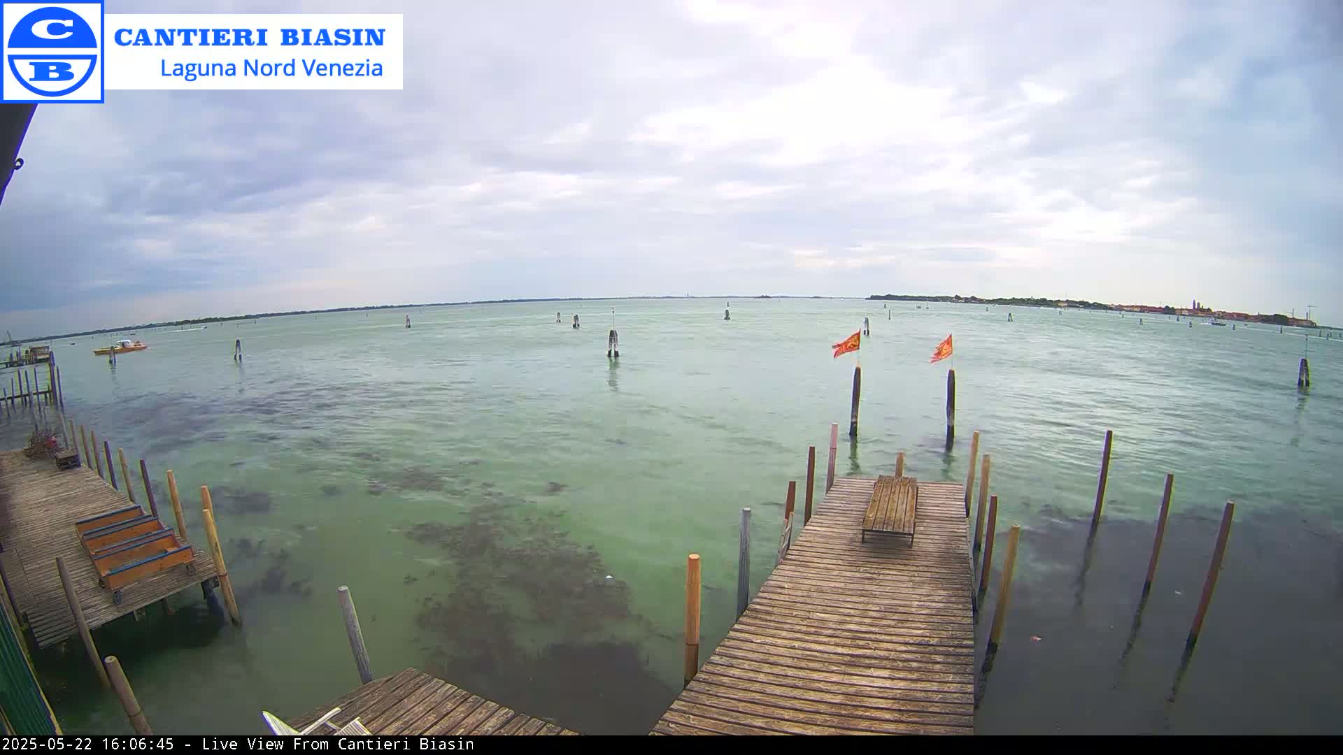 A wooden dock extends over calm, greenish water under a cloudy sky, with a few small boats and flags visible in the distance.