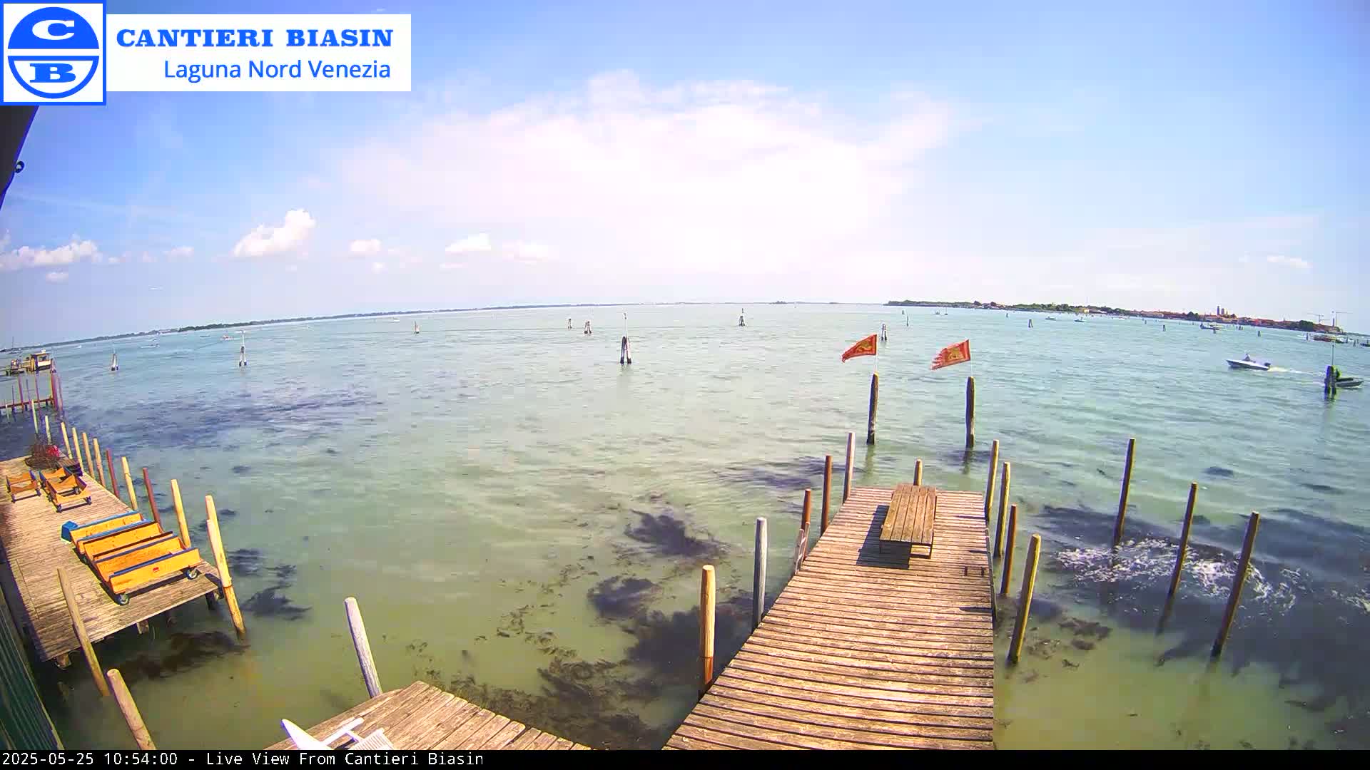 A wooden dock extends over shallow, calm, greenish water with a few boats and flags visible in the distance under a mostly sunny sky.