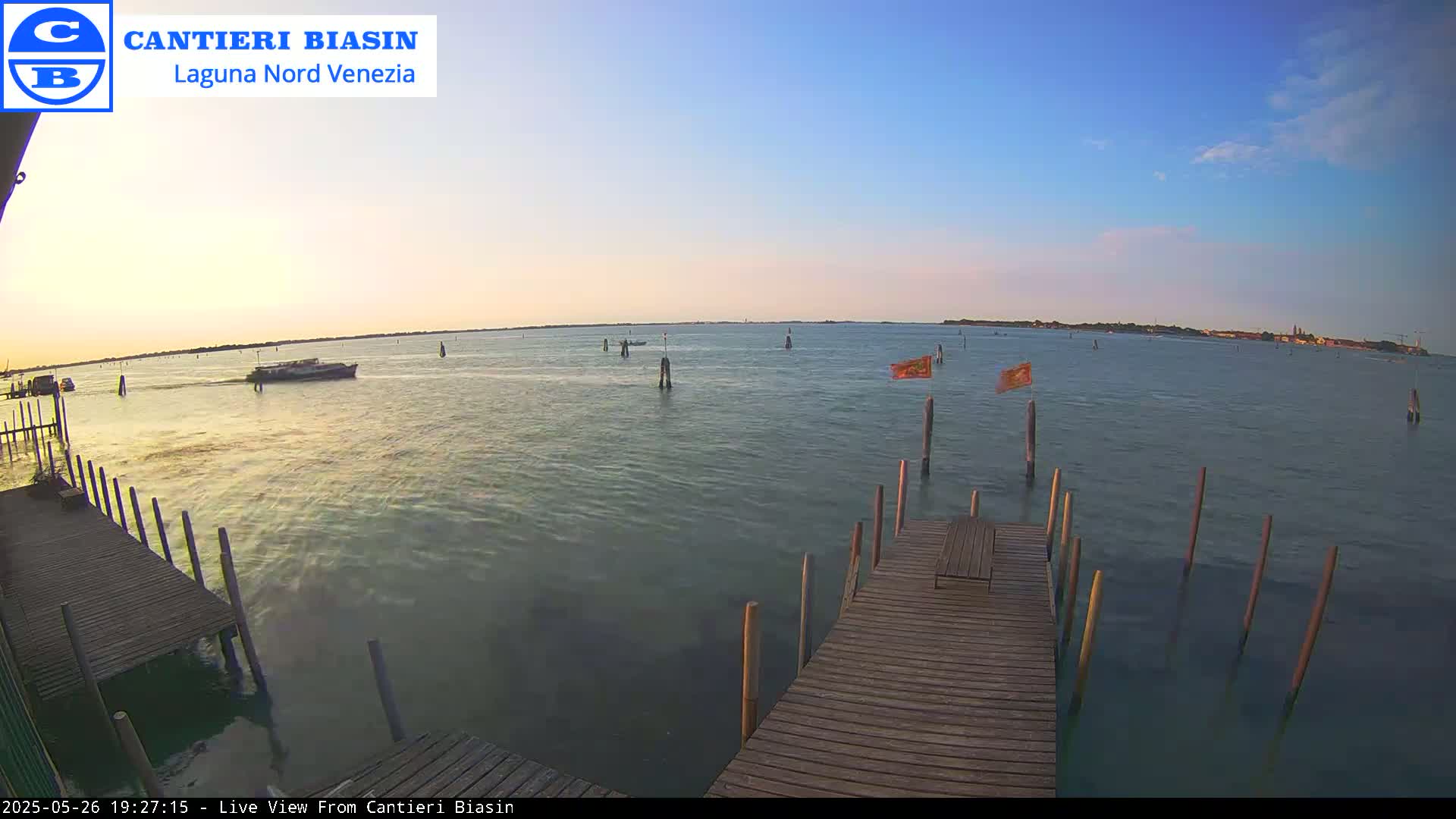 A calm body of water at sunset is viewed from a wooden dock, with several boats and posts visible in the distance.