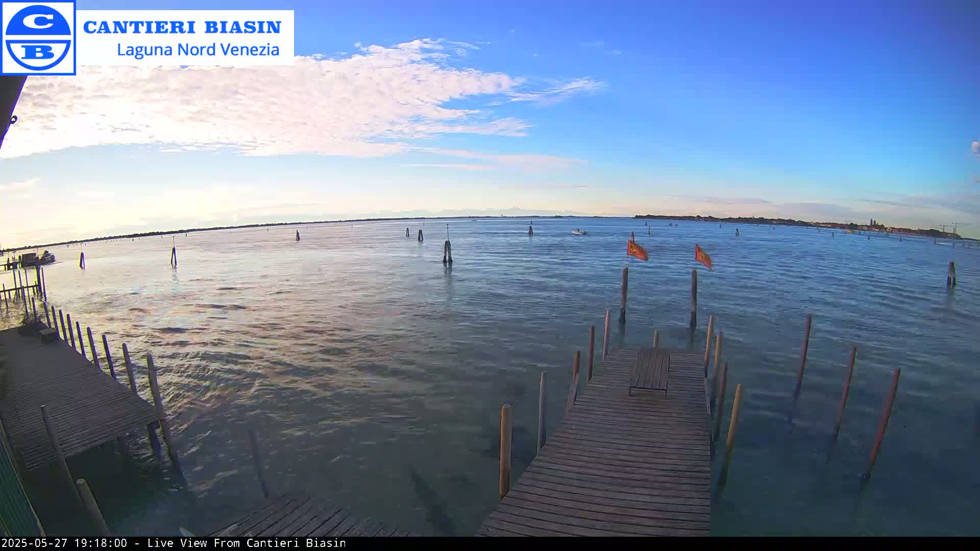 A wooden dock extends over calm, blue water under a mostly clear, light-blue sky, with a few boats and orange flags visible in the distance.