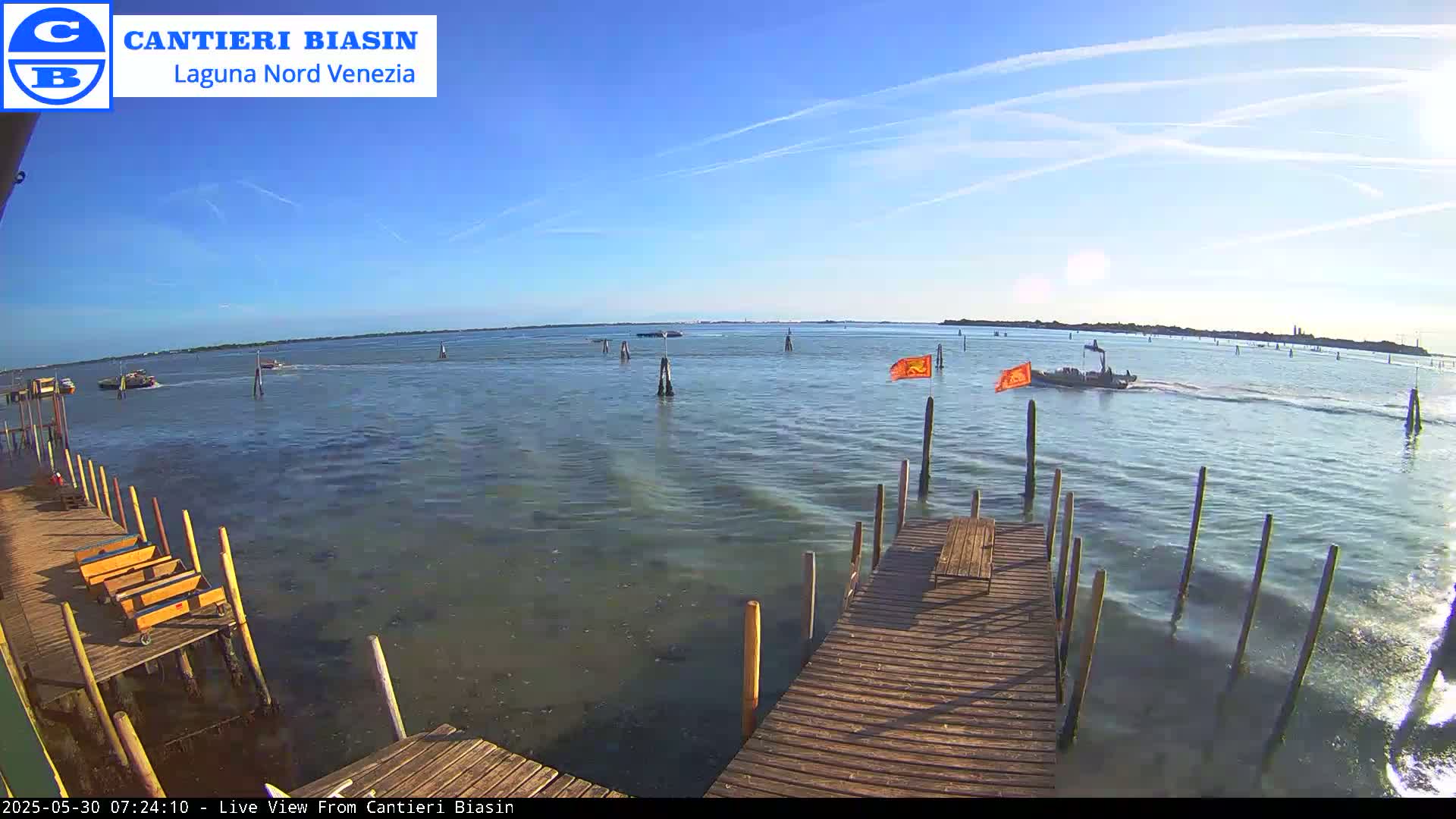 A wooden dock extends over calm, slightly rippled water, with several boats visible in the distance under a clear, sunny sky.