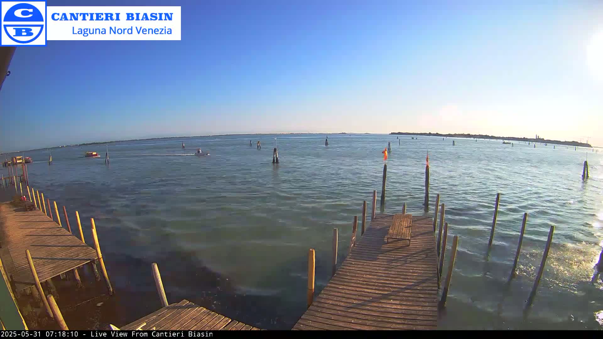 A wooden dock extends over calm, greenish-blue water under a clear blue sky, with several boats and markers visible in the distance.