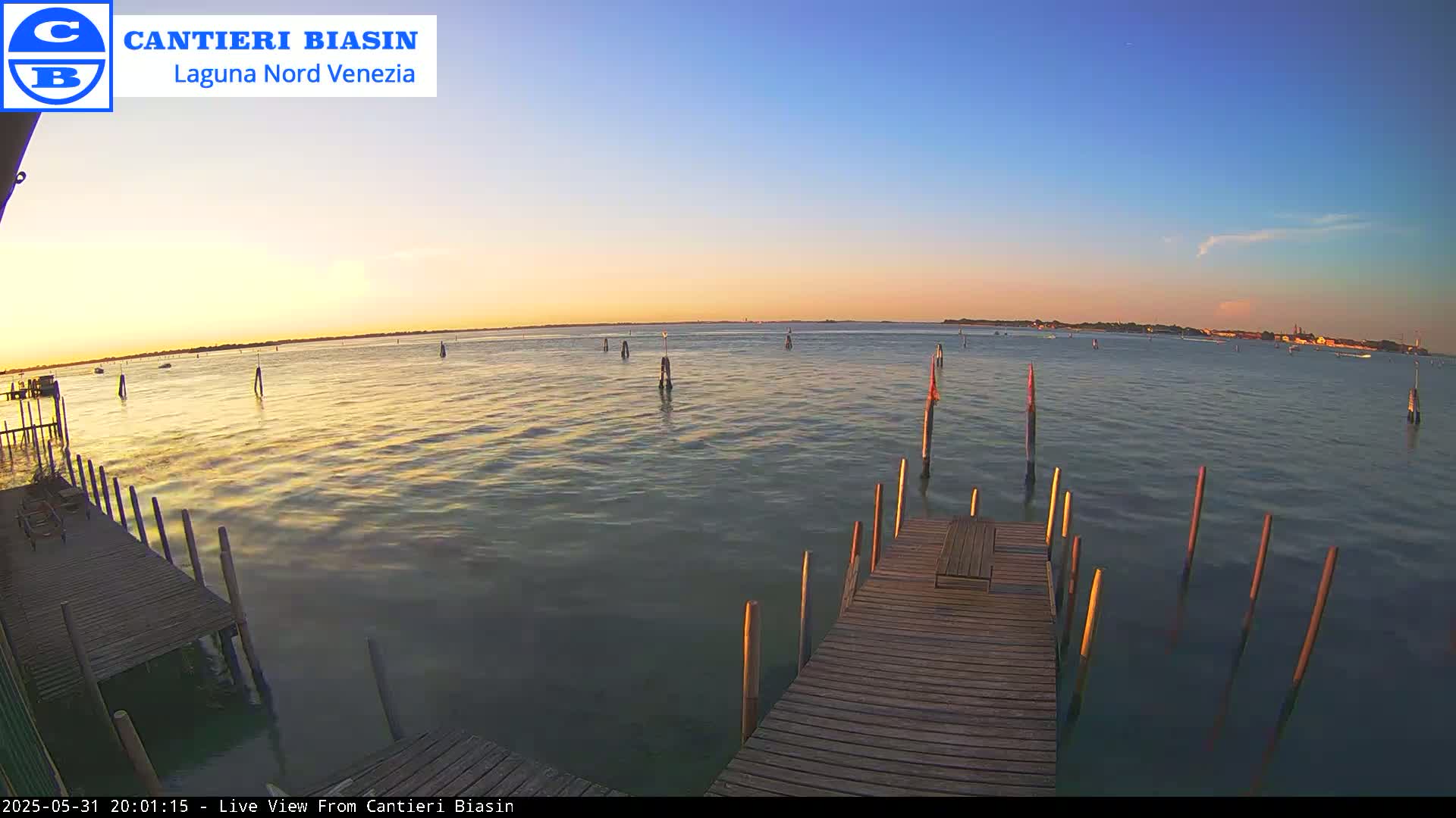 A wooden dock extends over calm water at sunset, with several posts in the water and land visible in the distance.