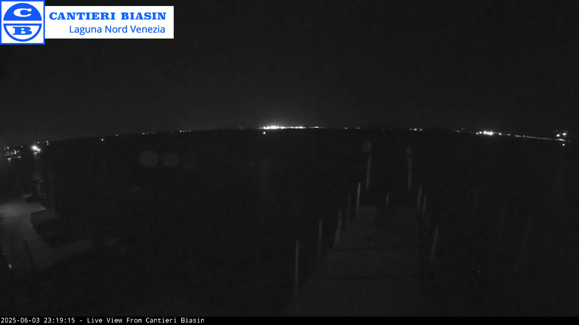 A nighttime, low-light view shows a dark pier extending into a dark body of water, with a distant line of lights visible on the horizon.
