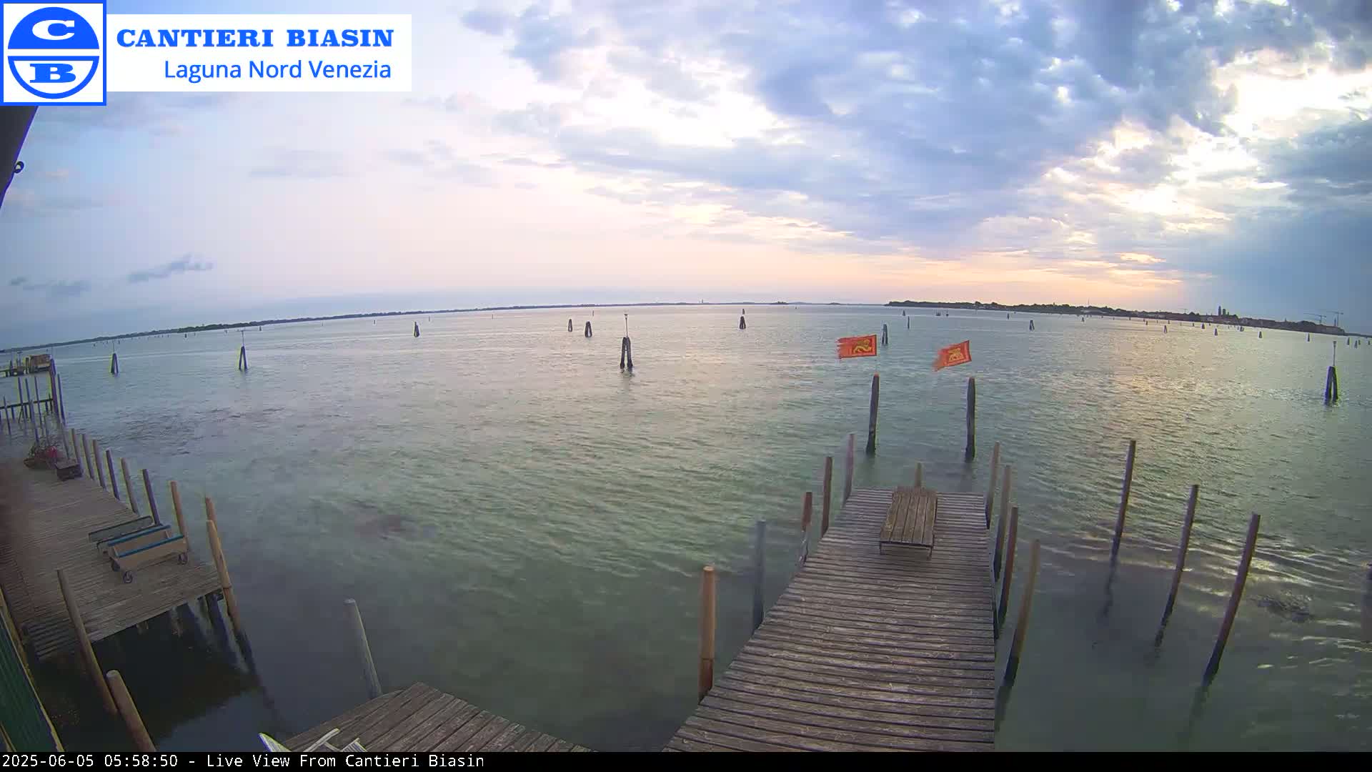 A wooden dock extends over calm water under a partly cloudy sky, with several posts and small orange flags visible in the water.