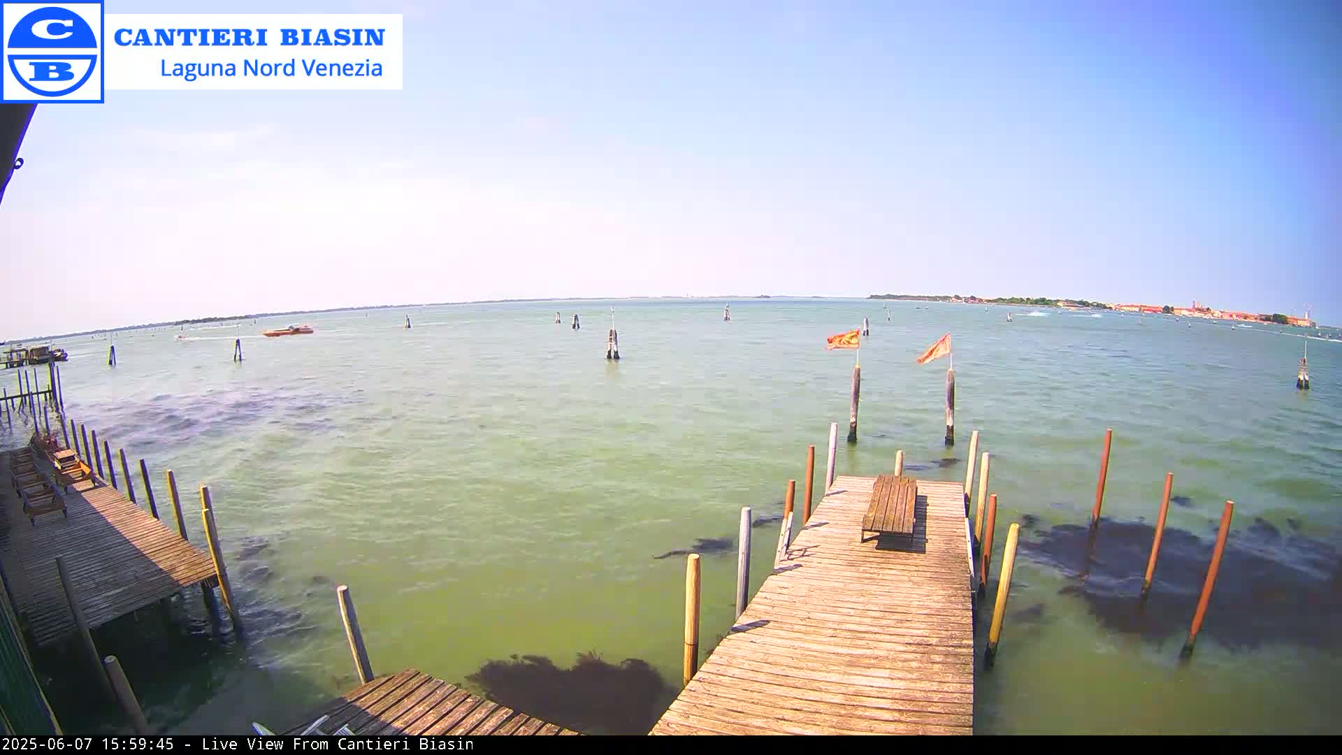 A wooden dock extends over calm, greenish water on a sunny day, with a few boats visible in the distance and land in the far background.
