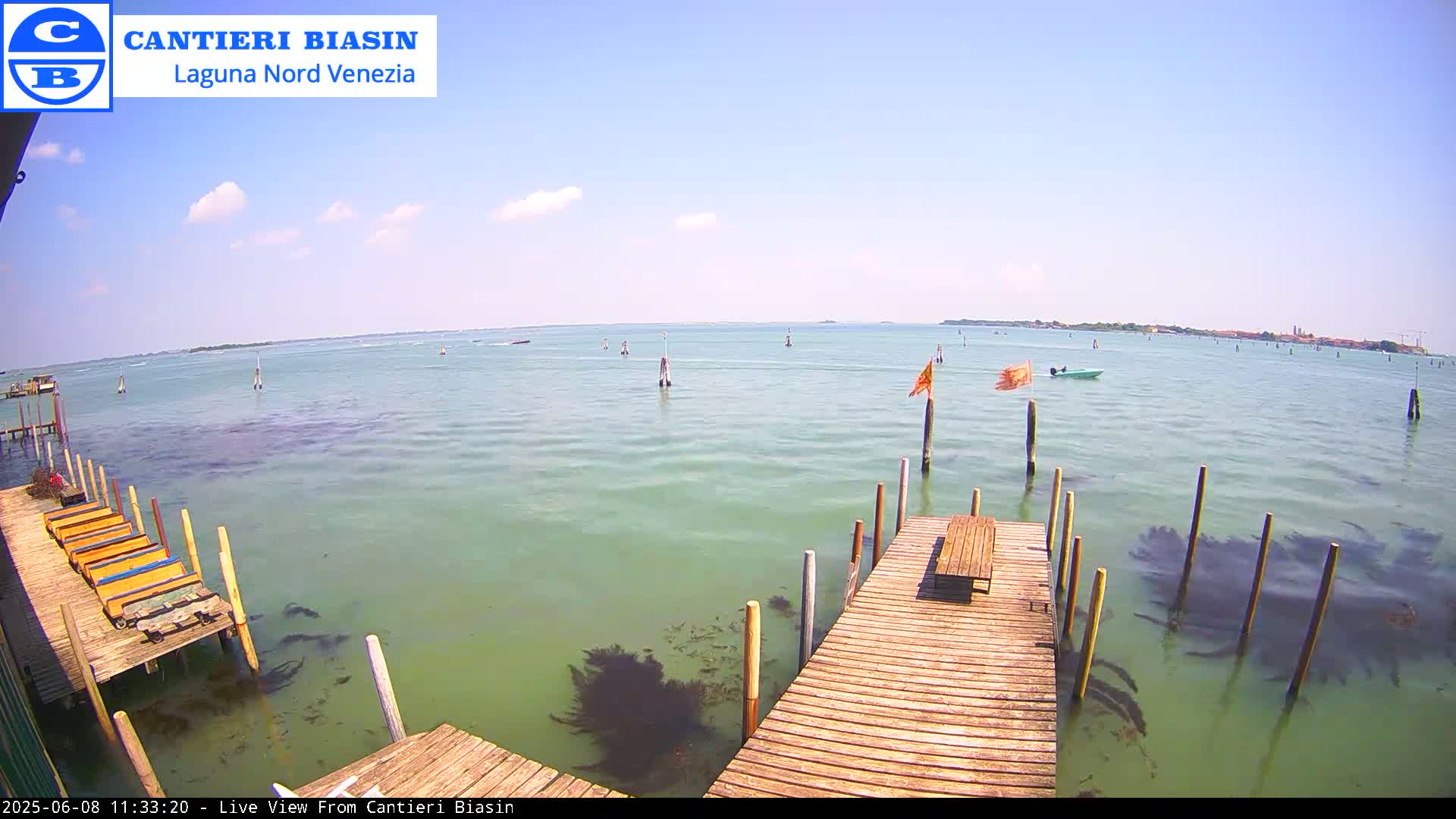 A calm, sunny day shows a wooden dock extending over light-green water, with several small boats visible in the distance and a landmass on the horizon.