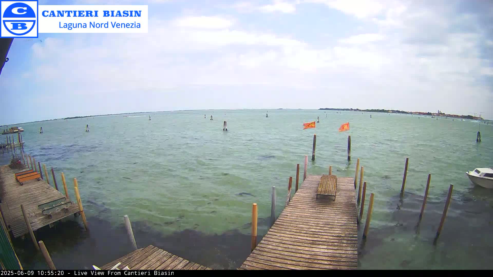 A partly cloudy day shows a view of calm, greenish water with a wooden dock extending into it; several poles stand in the water, and a small boat is visible in the distance.