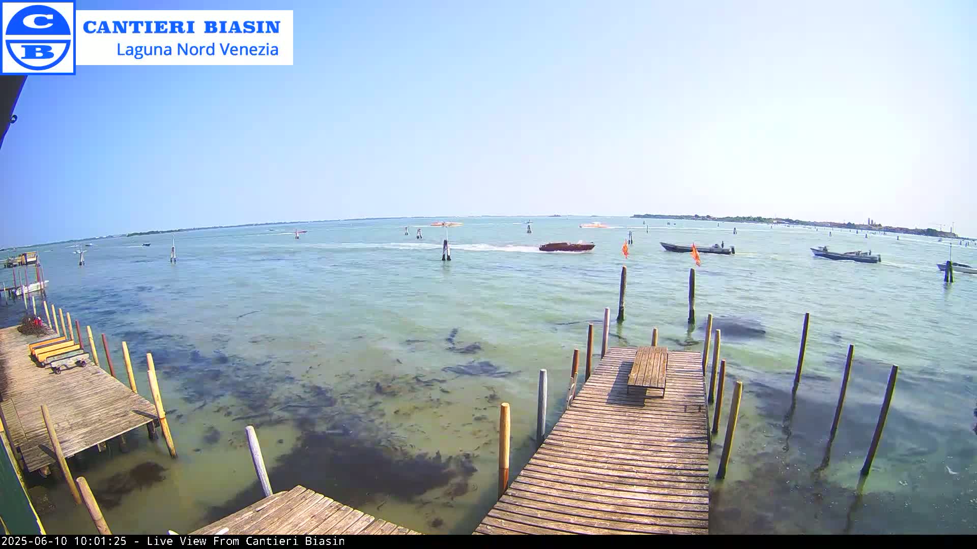 A wooden dock extends over calm, shallow, green-tinged water with several boats visible in the distance under a clear blue sky.