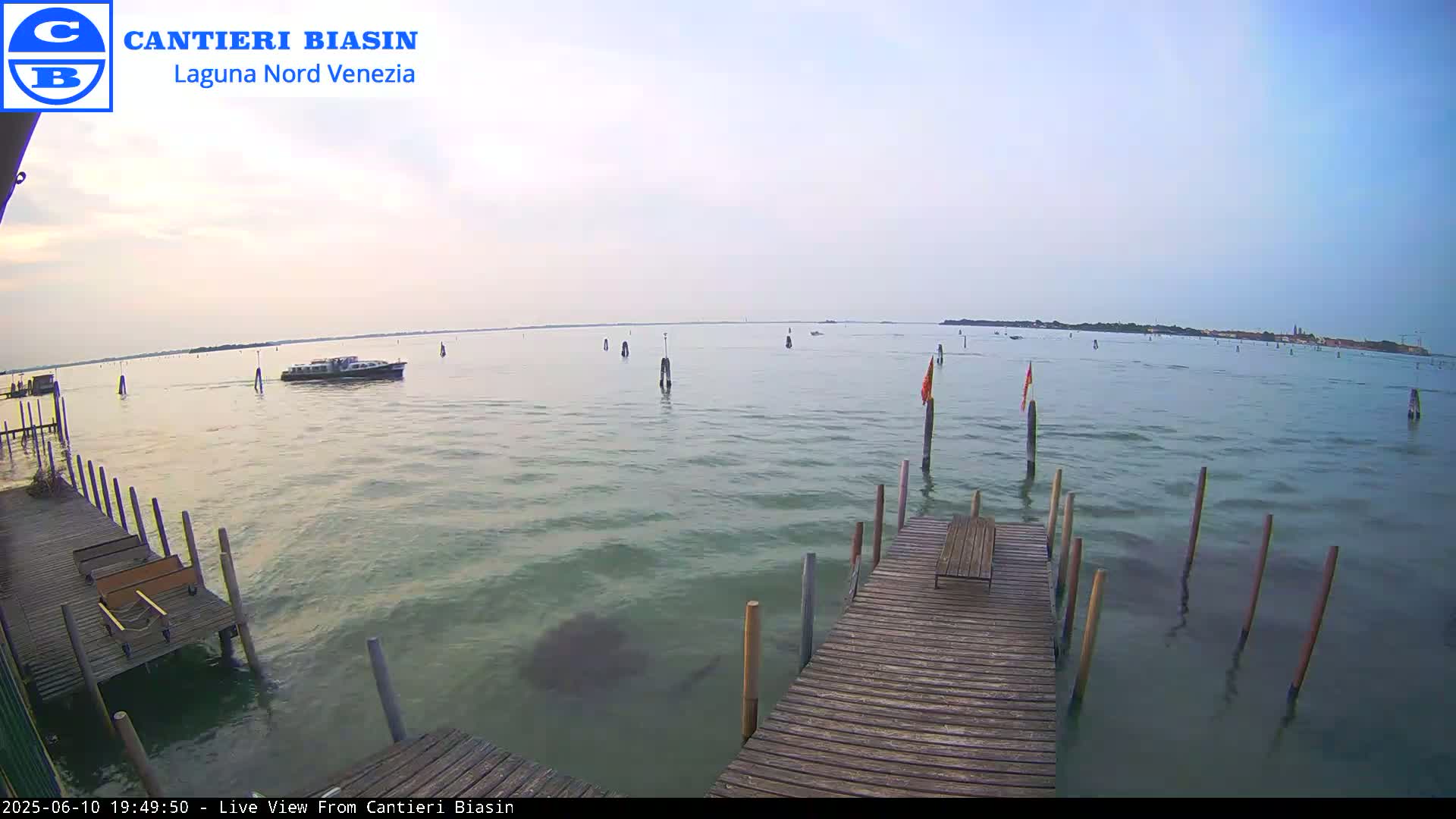 A calm body of water with a wooden dock, a boat in the distance, and a landmass visible on the horizon under a mostly clear sky.