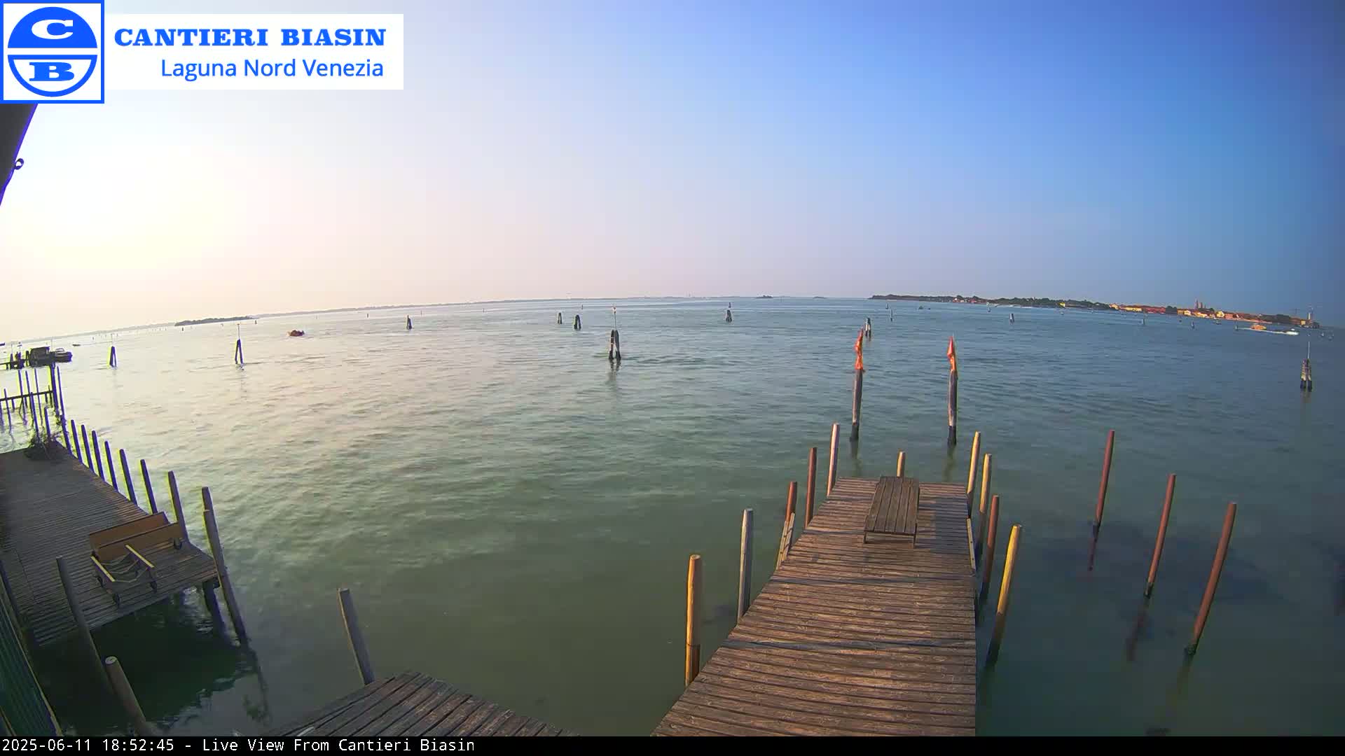 A wooden dock extends over calm, greenish water under a clear blue sky, with several posts in the water and land visible in the distance.