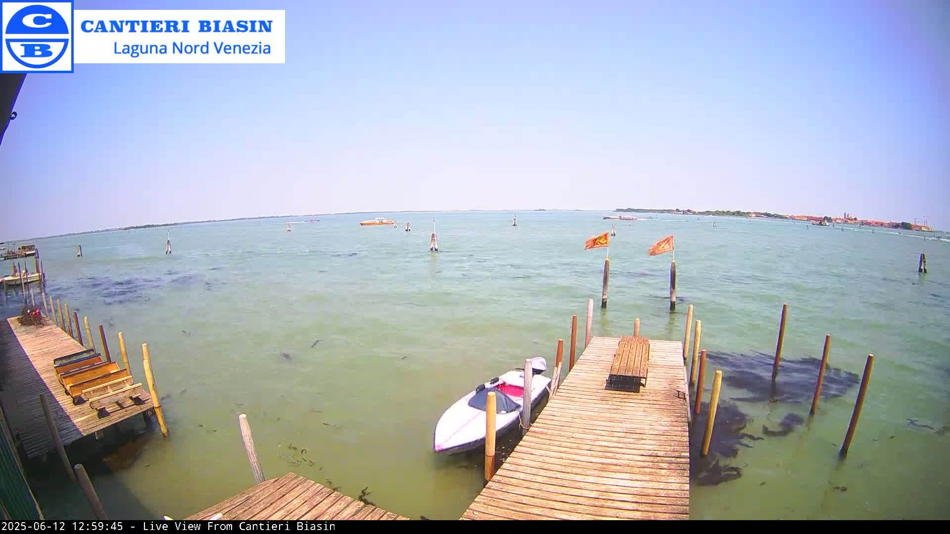 A calm, sunny day is shown overlooking a body of water with several boats and wooden docks, one of which has a small motorboat moored to it.