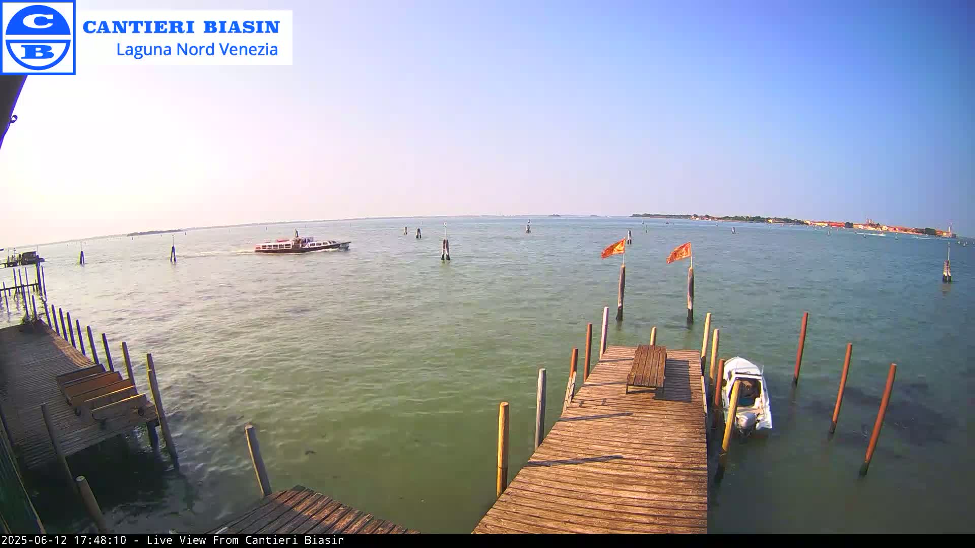 A wooden dock extends over calm, green-tinged water, with a small boat moored alongside and a larger vessel in the distance on a sunny day.