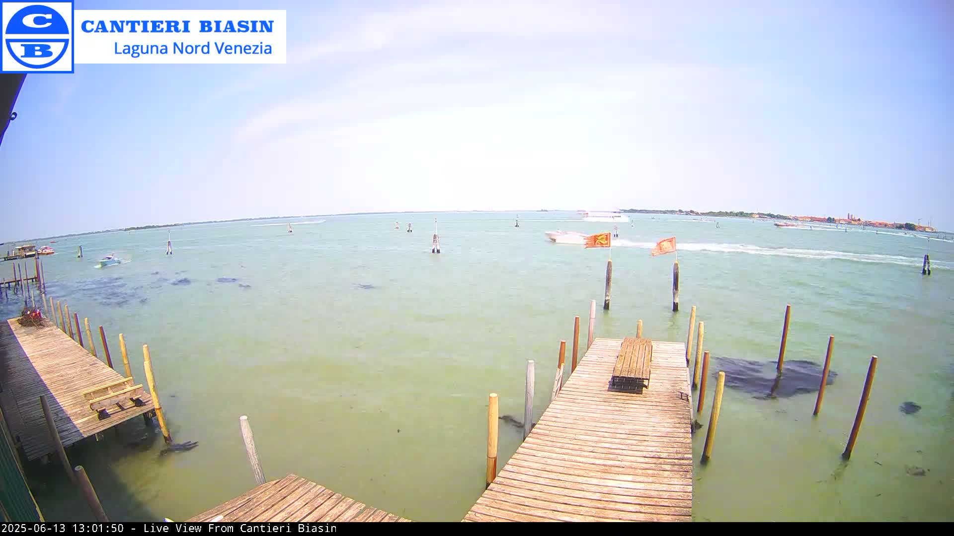 A wooden dock extends over shallow, light-green water, with several boats visible in the distance on a sunny day.