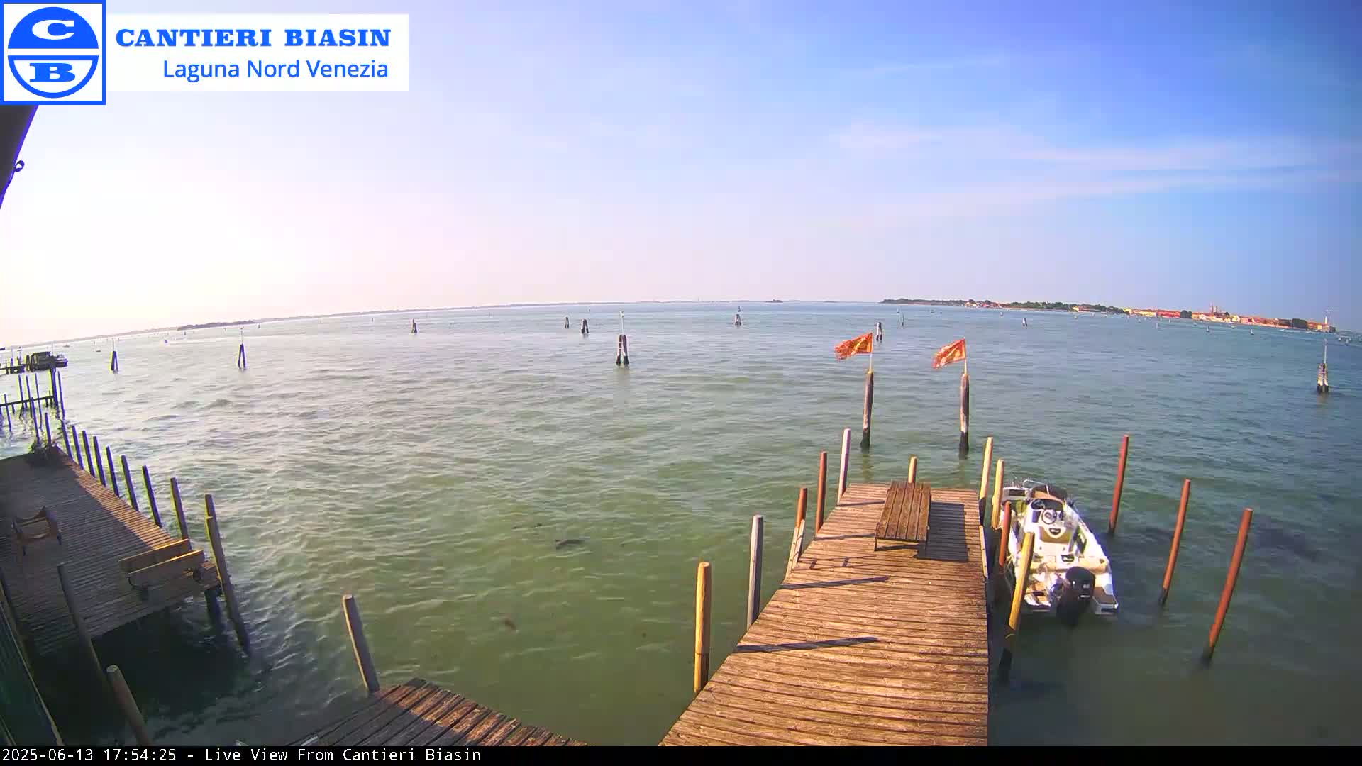 A wooden dock extends over calm, greenish water under a clear, sunny sky, with a small motorboat moored alongside and distant land visible on the horizon.