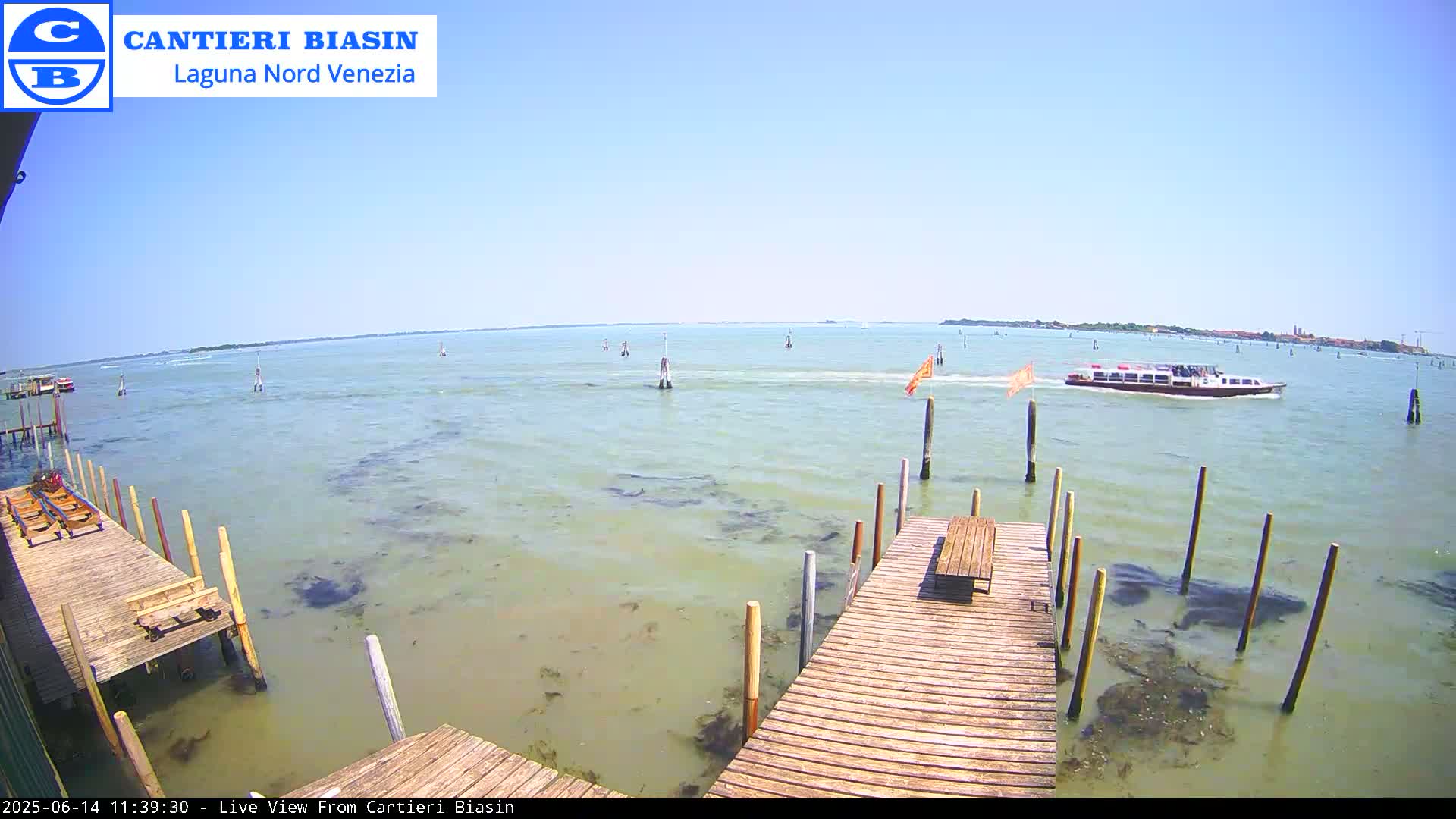A wooden dock extends over calm, light-green water on a sunny day, with a boat in the distance and several markers in the water.