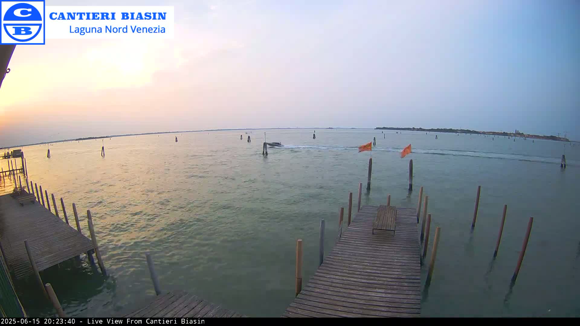 A calm body of water at sunset, featuring a wooden dock extending into the water, several posts in the water, and a small boat in the distance.