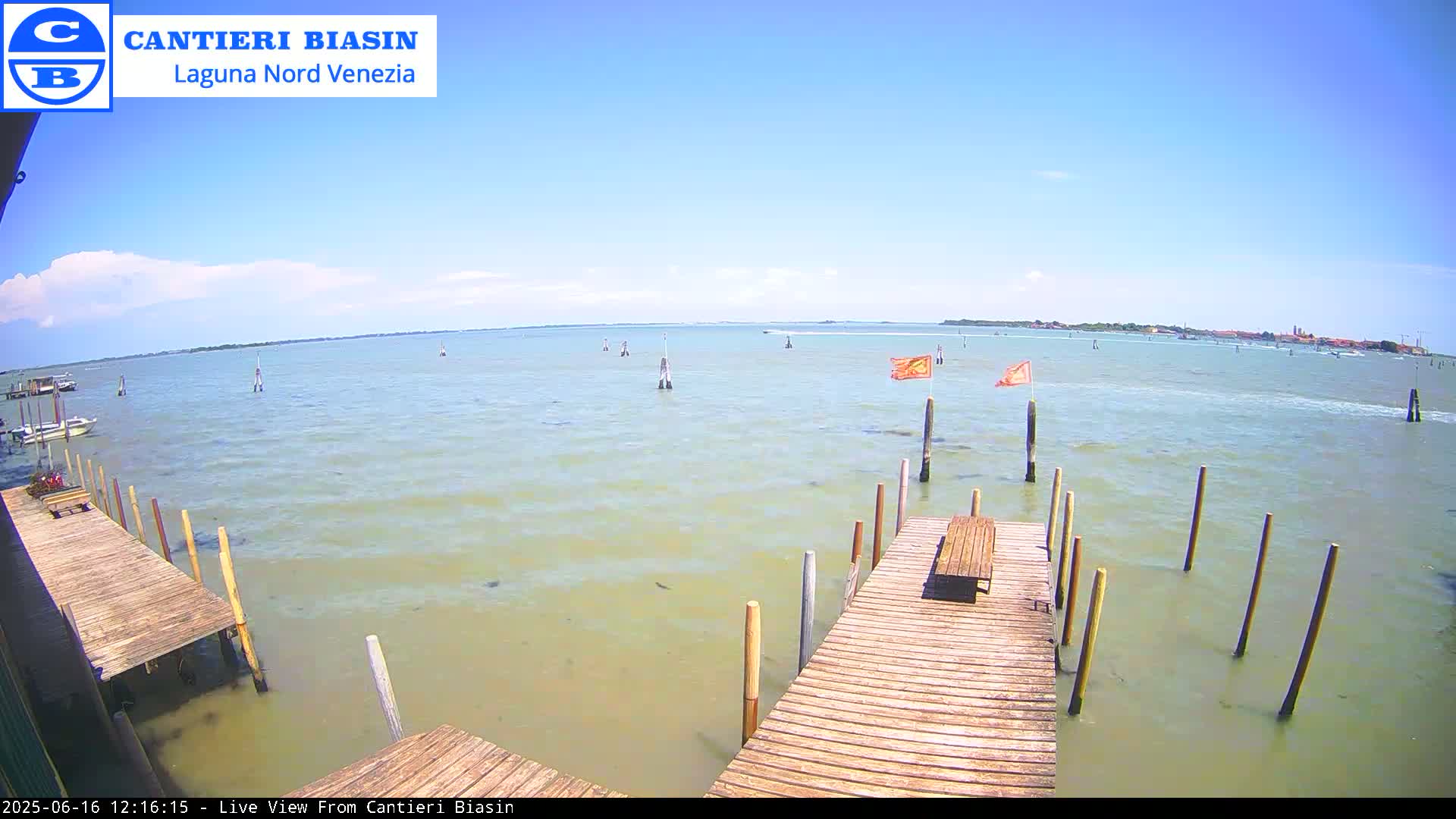 A wooden dock extends over calm, light-green water under a mostly clear, sunny sky; in the distance, land and some boats are visible.