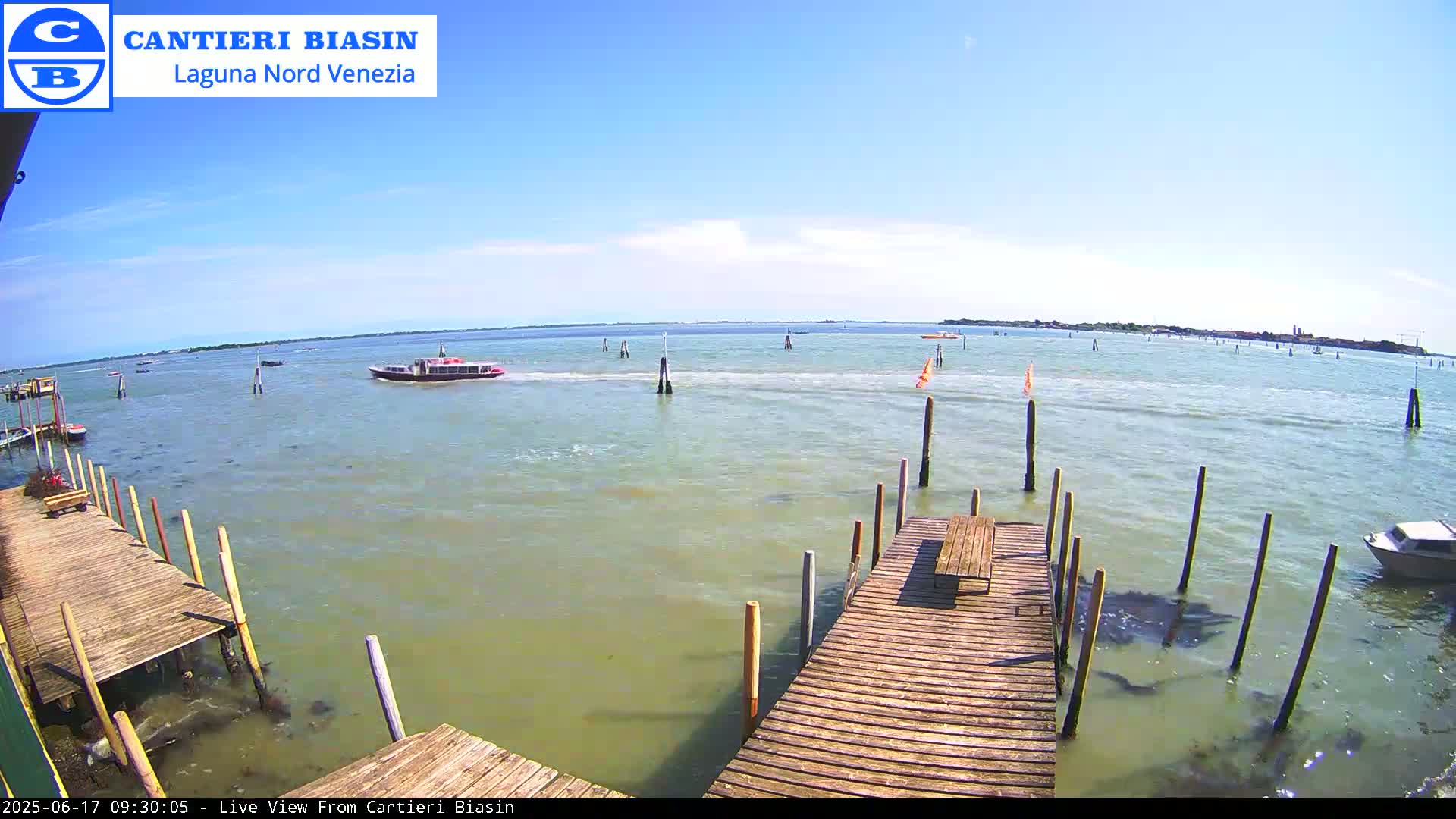 A calm, sunny day on a body of water shows a wooden dock with a picnic table, a small boat in the foreground, and a larger vessel further out on the water.