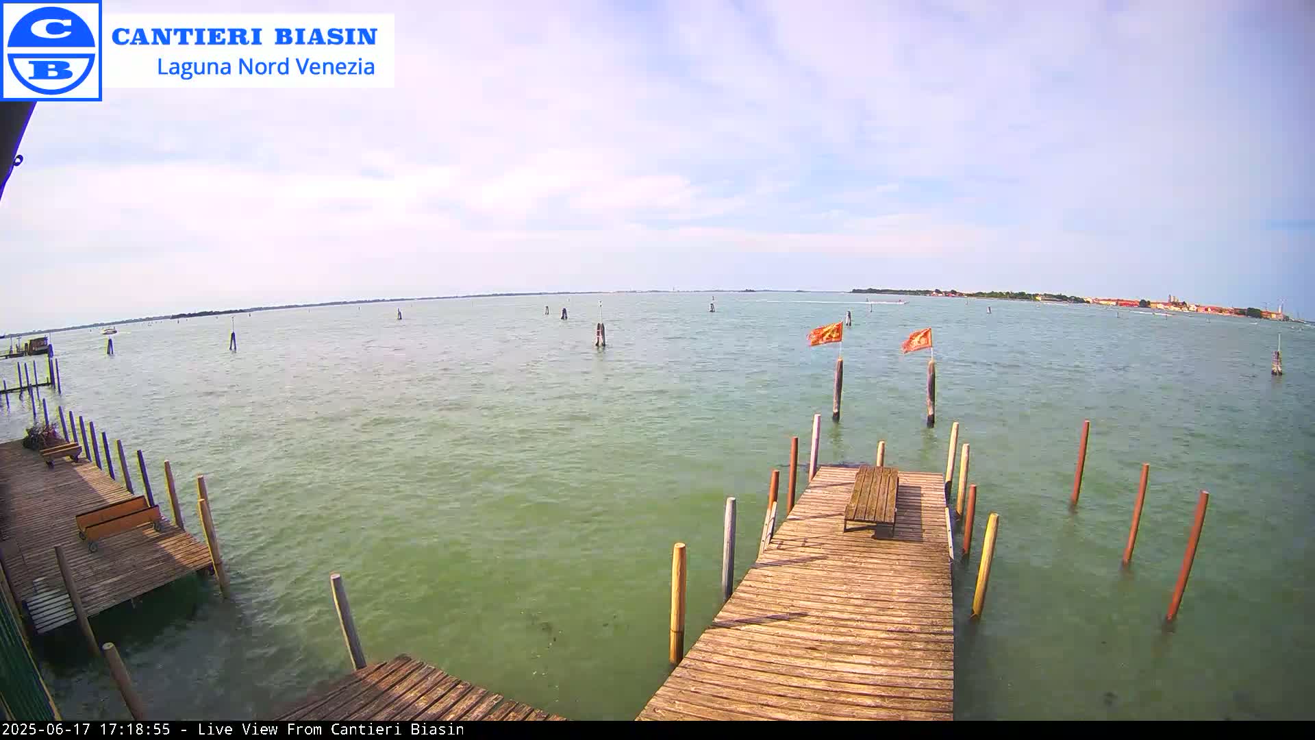 A wooden dock extends over calm, green water toward a distant landmass under a mostly clear sky.