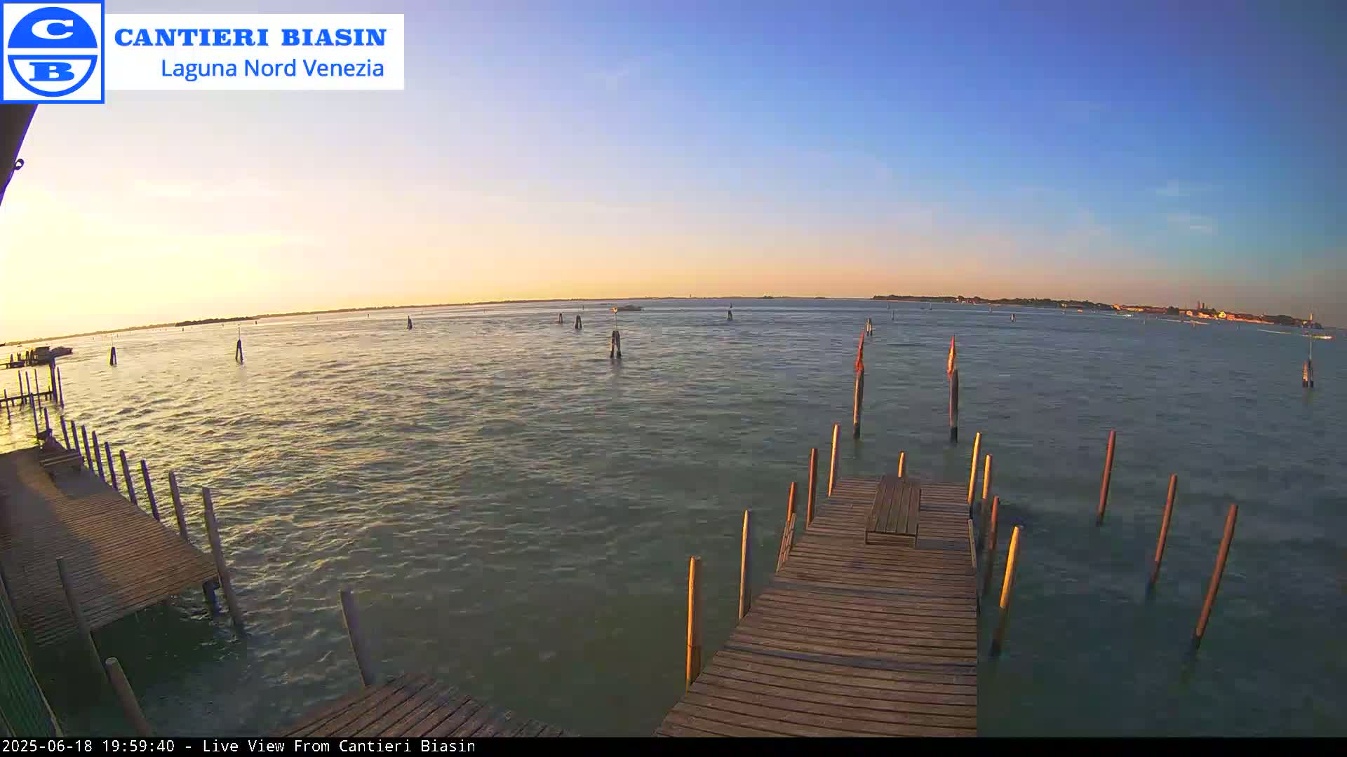 A wooden dock extends over calm water at sunset, with several posts in the water and land visible in the distance.
