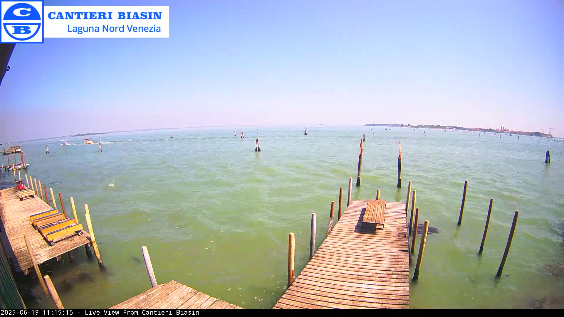 A wooden dock extends over calm, light green water, with several other docks and poles in the distance under a clear blue sky.