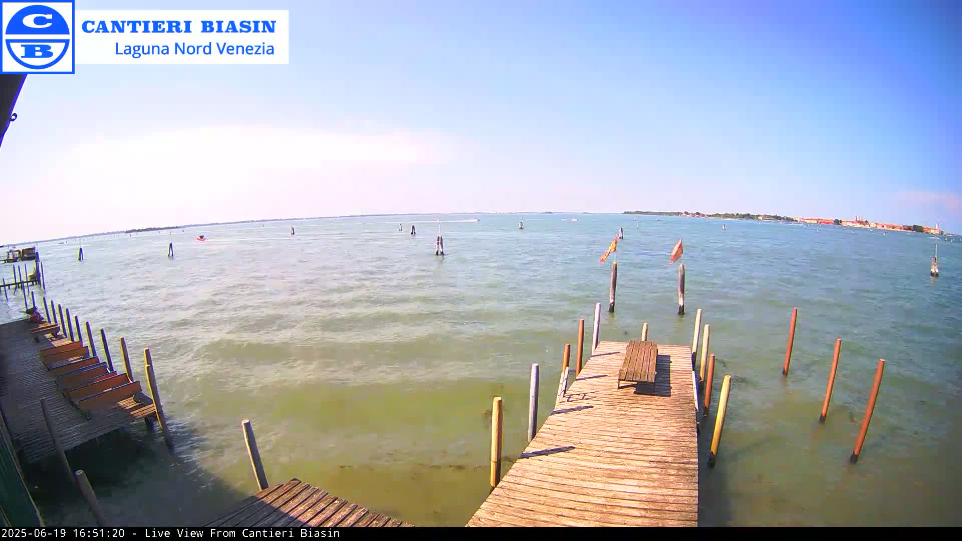 A wooden dock extends over a calm body of water on a sunny day, with a distant shoreline visible under a clear blue sky.