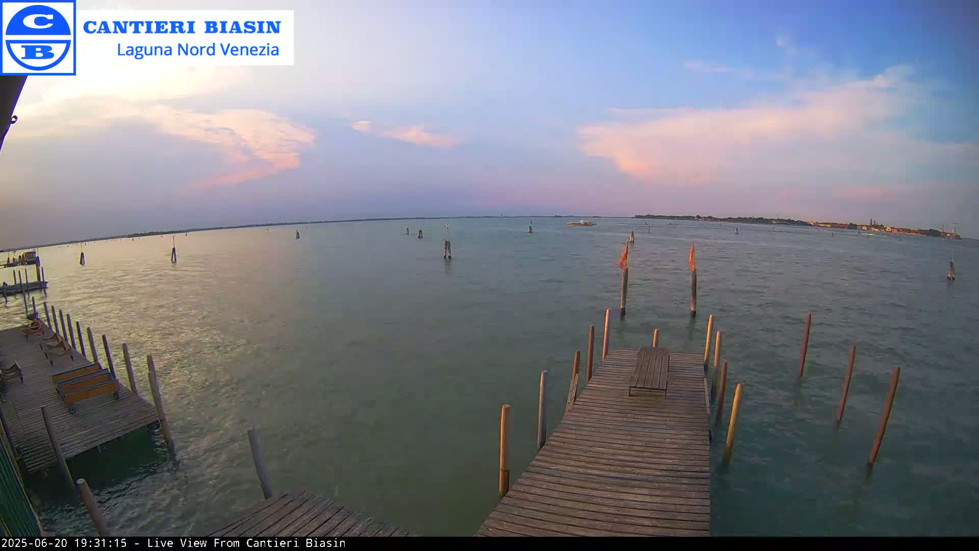 A wooden dock extends over calm water under a partly cloudy, pastel-colored sunset sky, with several posts and markers visible in the water.