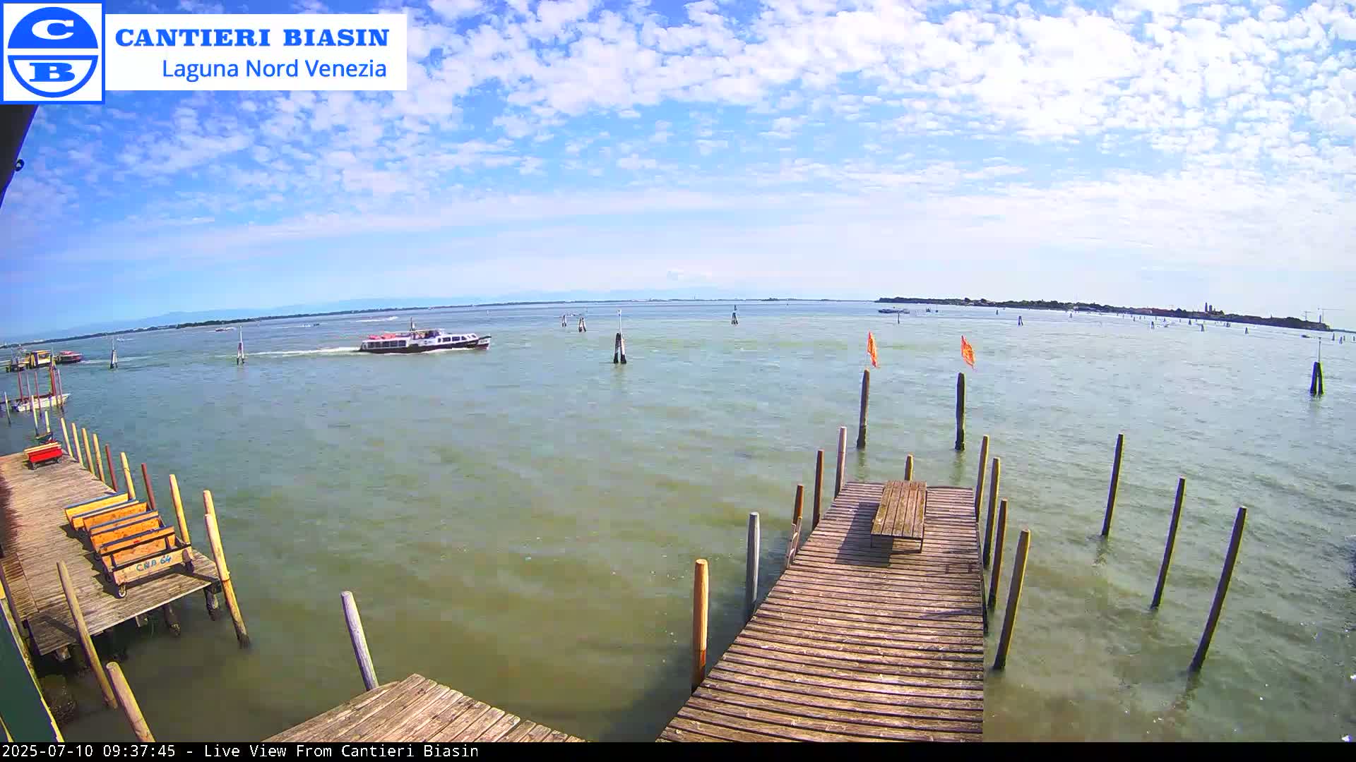 A partly cloudy day on a calm body of water shows boats passing by a wooden dock with a bench.