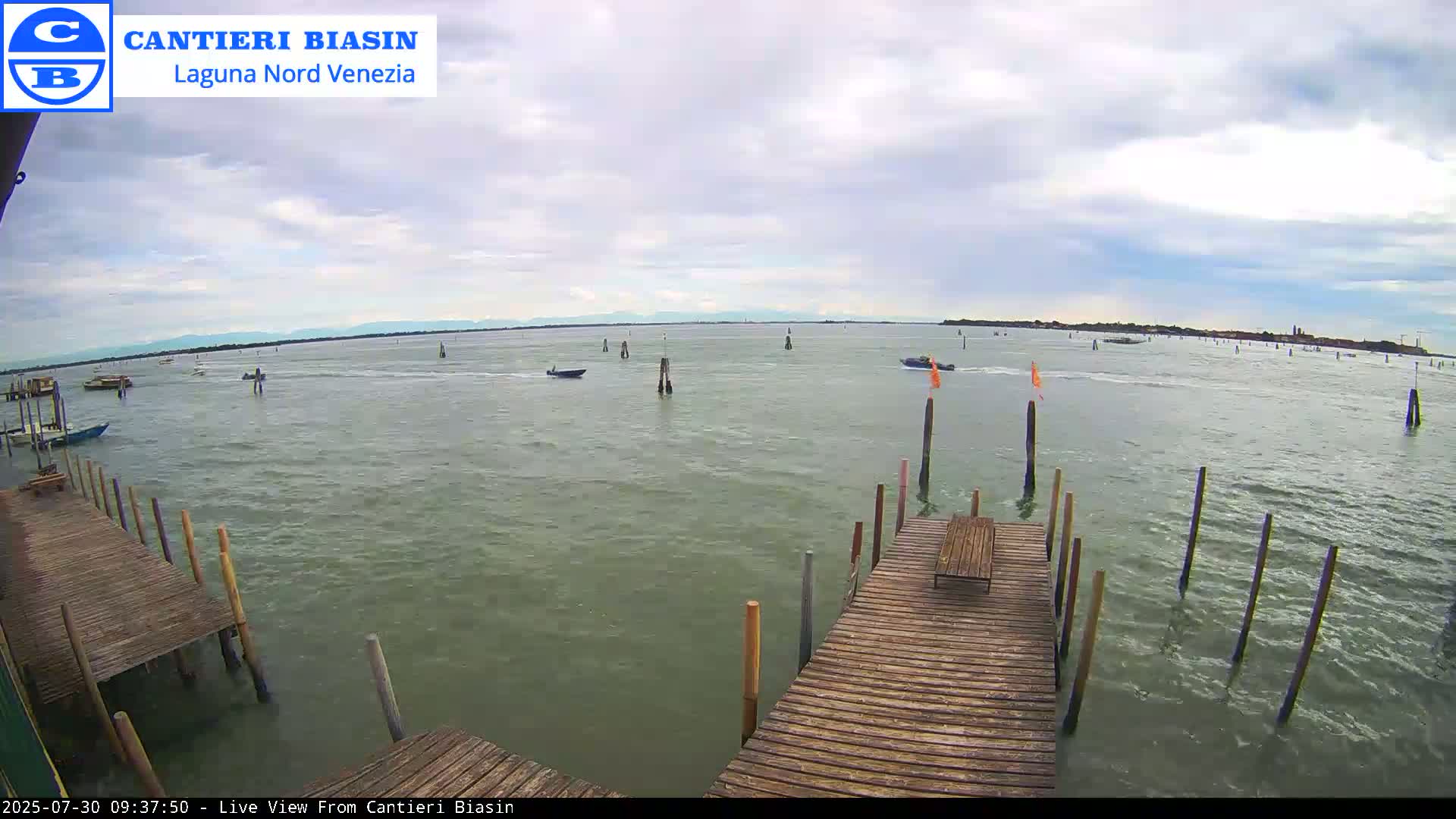 A wooden dock extends over calm, green water dotted with boats and wooden posts under a partly cloudy sky.