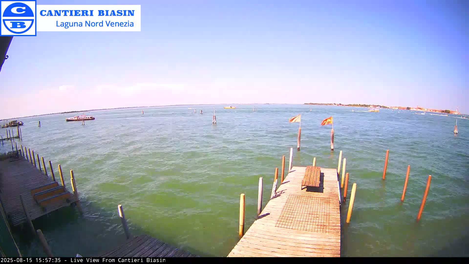 A wooden dock extends over calm, green water dotted with boats and wooden posts under a partly cloudy sky.